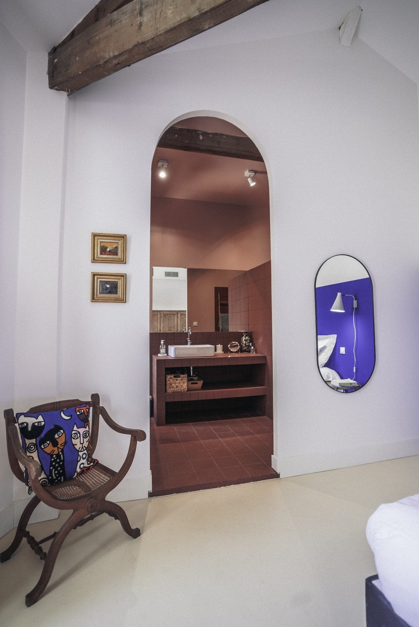 A cozy room features a wooden chair with vibrant patterned cushions, an oval mirror on the wall, and a view into a bathroom with pink walls and fixtures. The room has exposed wooden beams and soft, neutral-colored walls. Two small framed artworks adorn the wall.