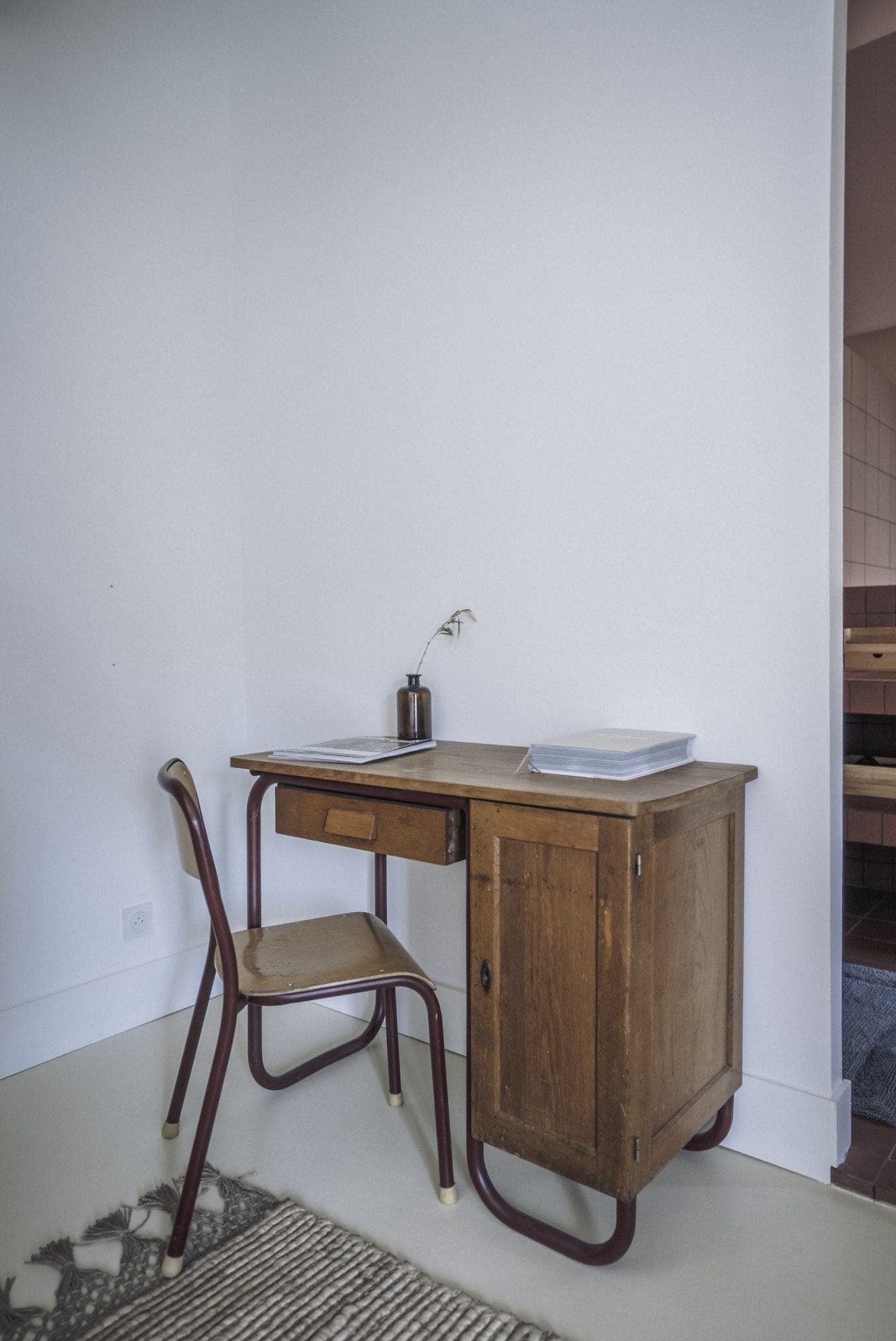 Un coin bureau minimaliste comprend un bureau en bois vintage avec un petit tiroir et une armoire d'un côté. Une chaise simple avec un siège et un dossier en bois est positionnée au bureau. Une plante dans un petit vase et deux cahiers sont placés sur le bureau. Un tapis tissé est partiellement visible sur le sol.