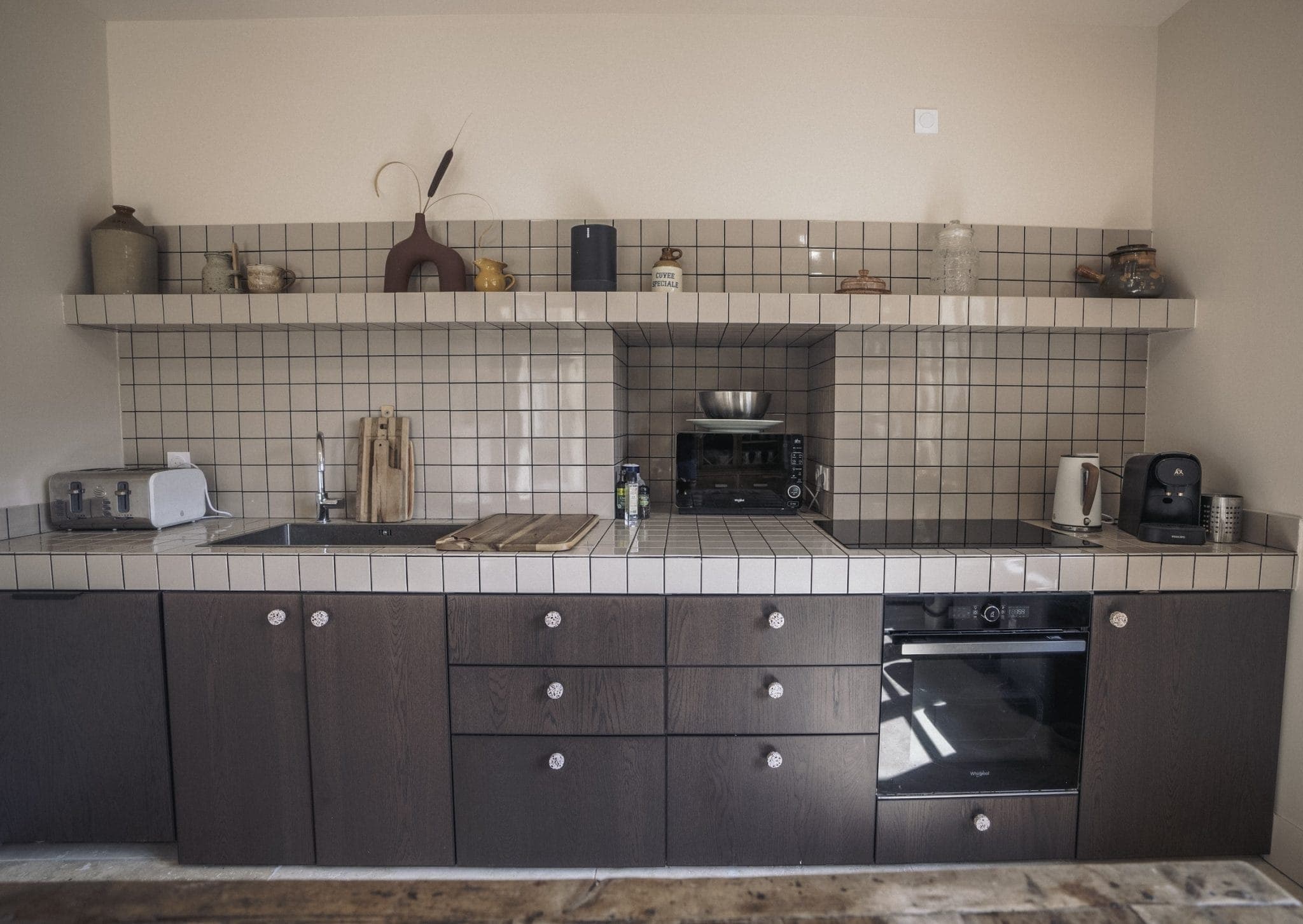 A kitchen with tiled walls and a wooden countertop featuring various appliances such as a toaster, coffee maker, oven, and kettle. Shelves above hold jars and decorative items, while a wooden table is partially visible in the foreground.