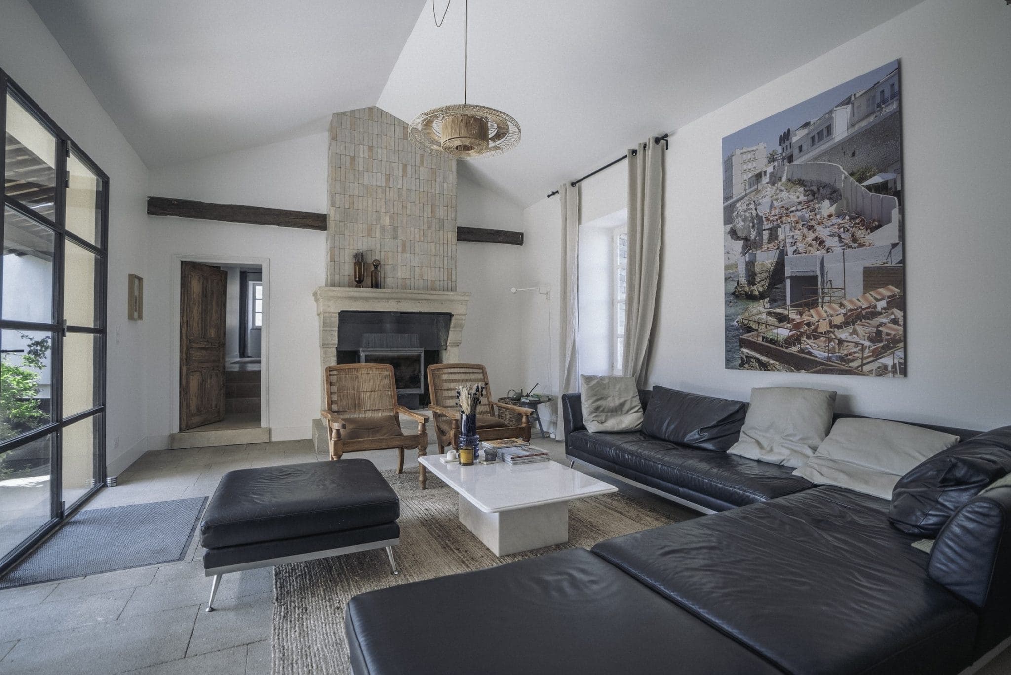A modern living room featuring a black leather sectional sofa, two wicker chairs, and a stone fireplace. A large photo hangs on the wall above the sofa, and a woven pendant light is suspended from the ceiling. Large glass doors provide natural light.