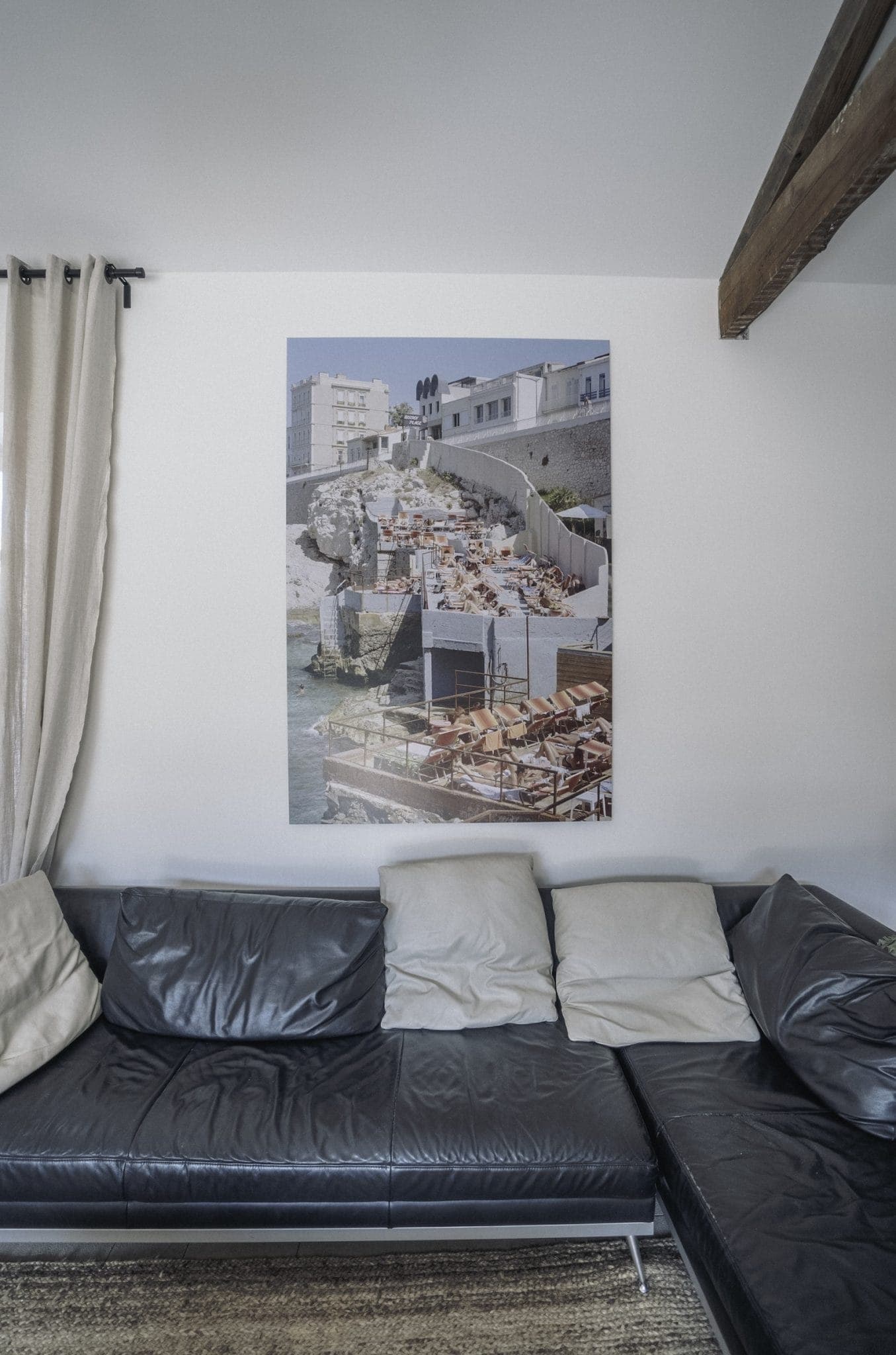 A modern living room with a dark leather sectional sofa adorned with light-colored pillows. On the white wall above the sofa hangs a large photograph of a coastal scene with buildings and beach chairs. To the left, a beige curtain hangs from a black curtain rod.