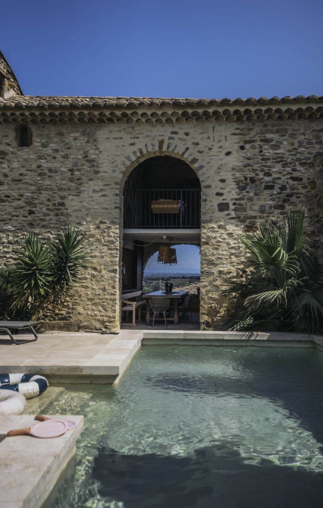 A serene outdoor pool reflects a stone building with an archway that frames a distant ocean view. The pool area is surrounded by lush green plants and leads to a shaded patio with seating. The sky above is clear and blue.