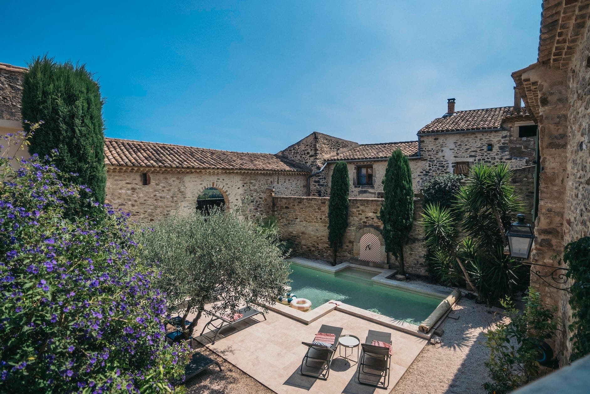 A rustic stone courtyard with a small, rectangular pool surrounded by lush plants and trees. Two lounge chairs sit on a patio, and the backdrop features historic stone buildings with tiled roofs under a clear blue sky.