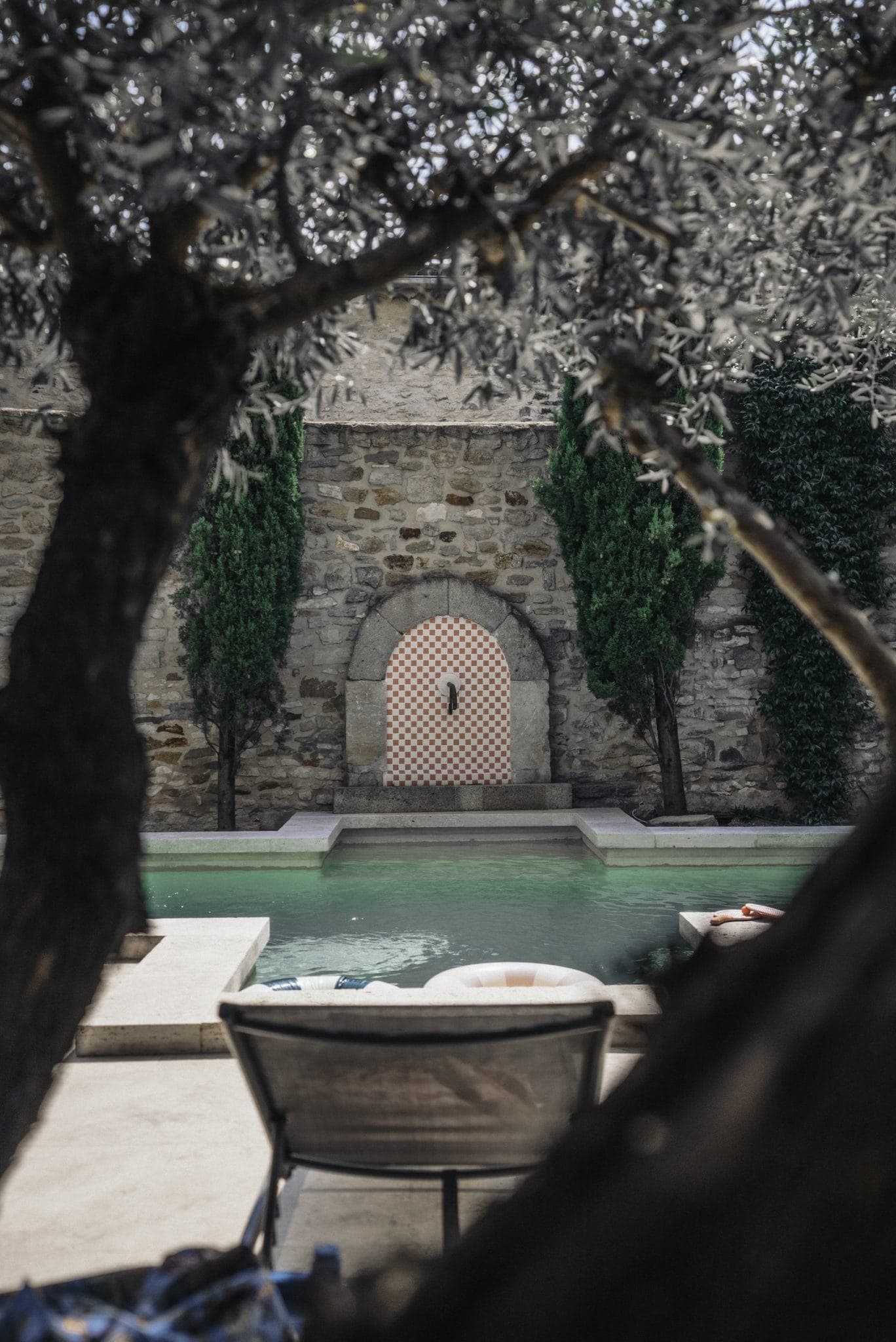 A serene pool area with a lounging chair in the foreground, framed by tree branches. The pool has a greenish hue and leads up to a stone wall adorned with a patterned archway and flanked by tall green bushes.