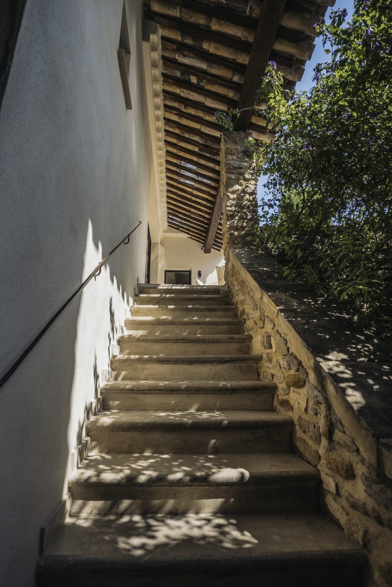 A stone staircase leads up to a doorway under a wooden beamed roof. The right side of the staircase is bordered by a stone wall, while the left side features a smooth plastered wall with a handrail. Foliage casts dappled shadows on the steps in bright sunlight.