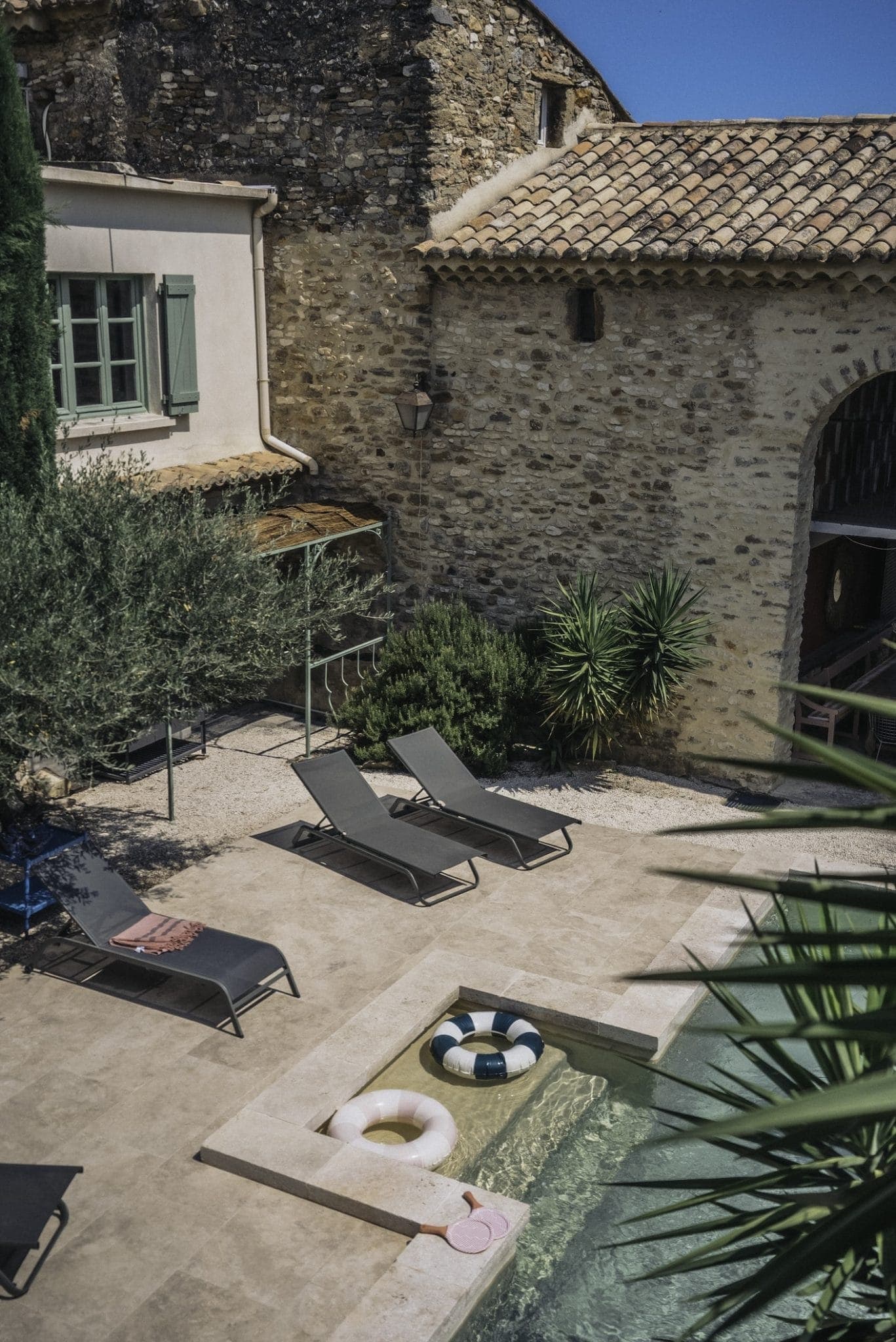 A serene courtyard featuring a small square pool with inflatable pool floats, surrounded by four sun loungers with towels. The area is flanked by stone buildings with terracotta roofs and lush green plants, against a backdrop of a clear blue sky.