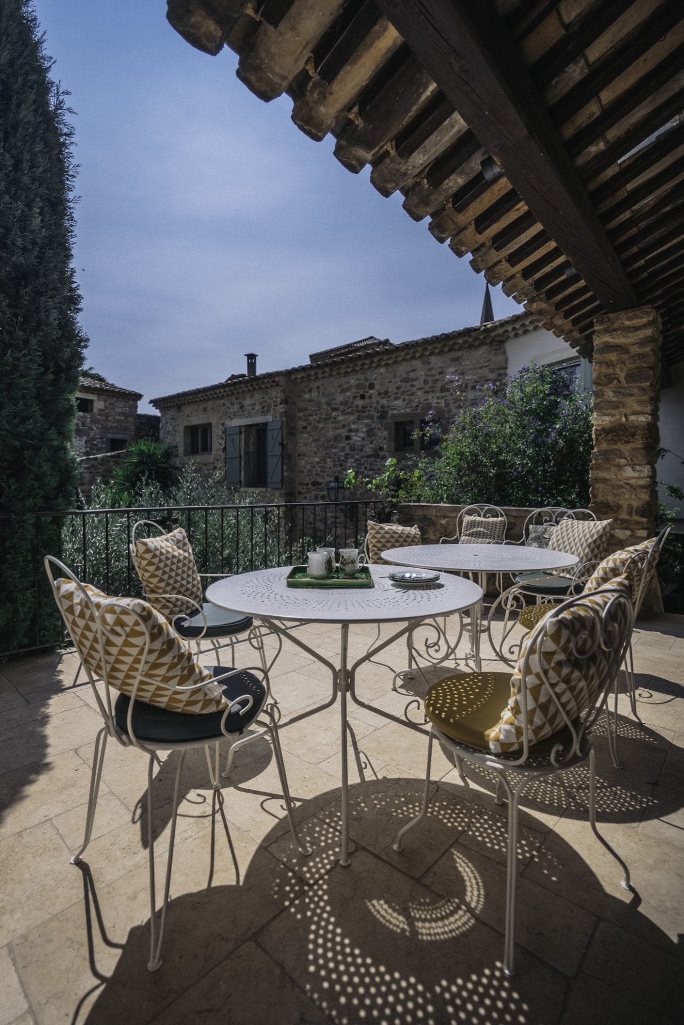 An outdoor patio with a white metal table and chairs adorned with yellow and white cushioned pillows under a rustic wooden pergola. The area is surrounded by stone buildings and lush greenery, creating a tranquil and inviting atmosphere. The sky is clear and blue.