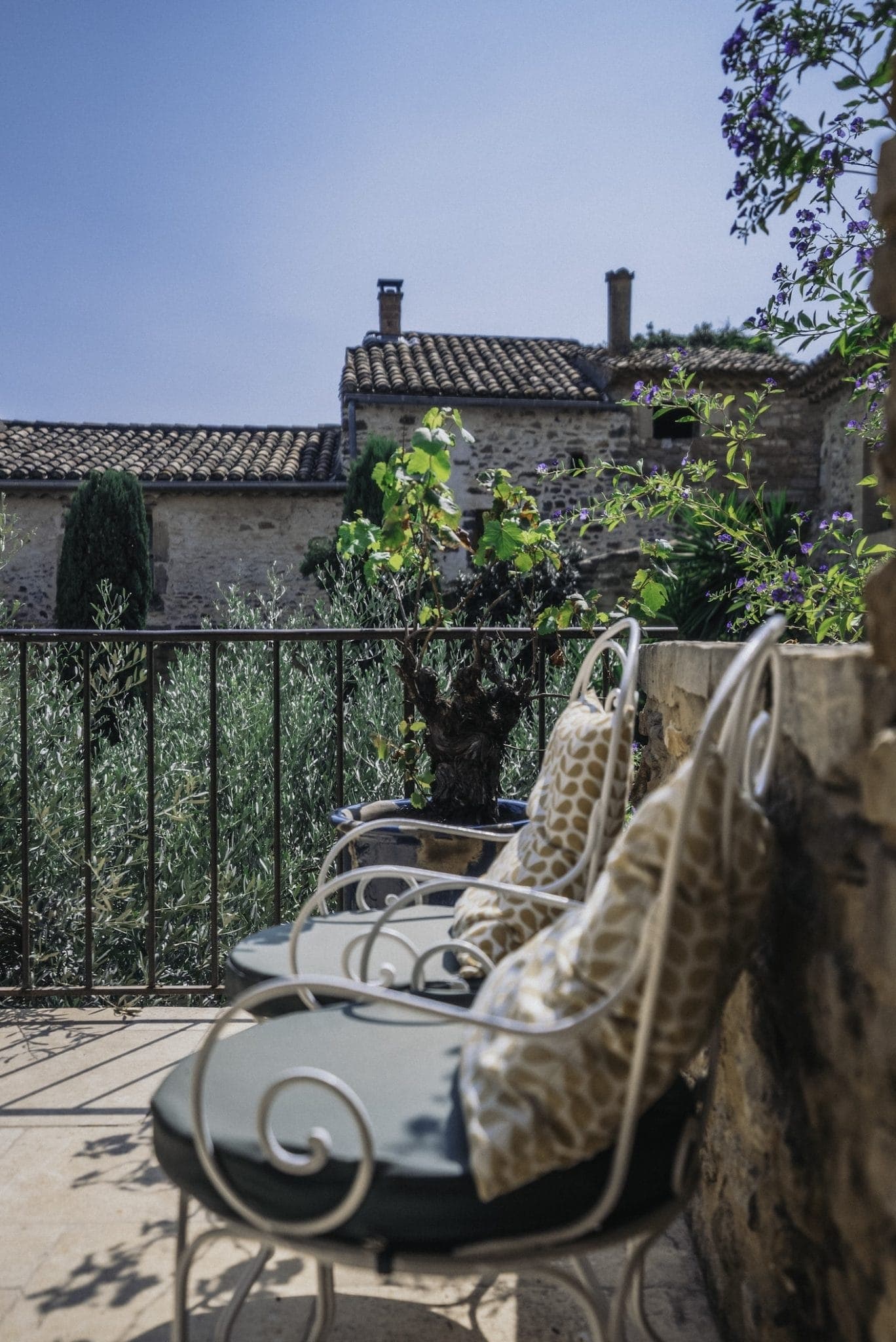 A sunny patio with two white wrought-iron chairs featuring green cushions and patterned throw pillows sits beside a stone wall adorned with a small shrub. In the background, there are rustic stone buildings with tiled roofs and an olive grove.