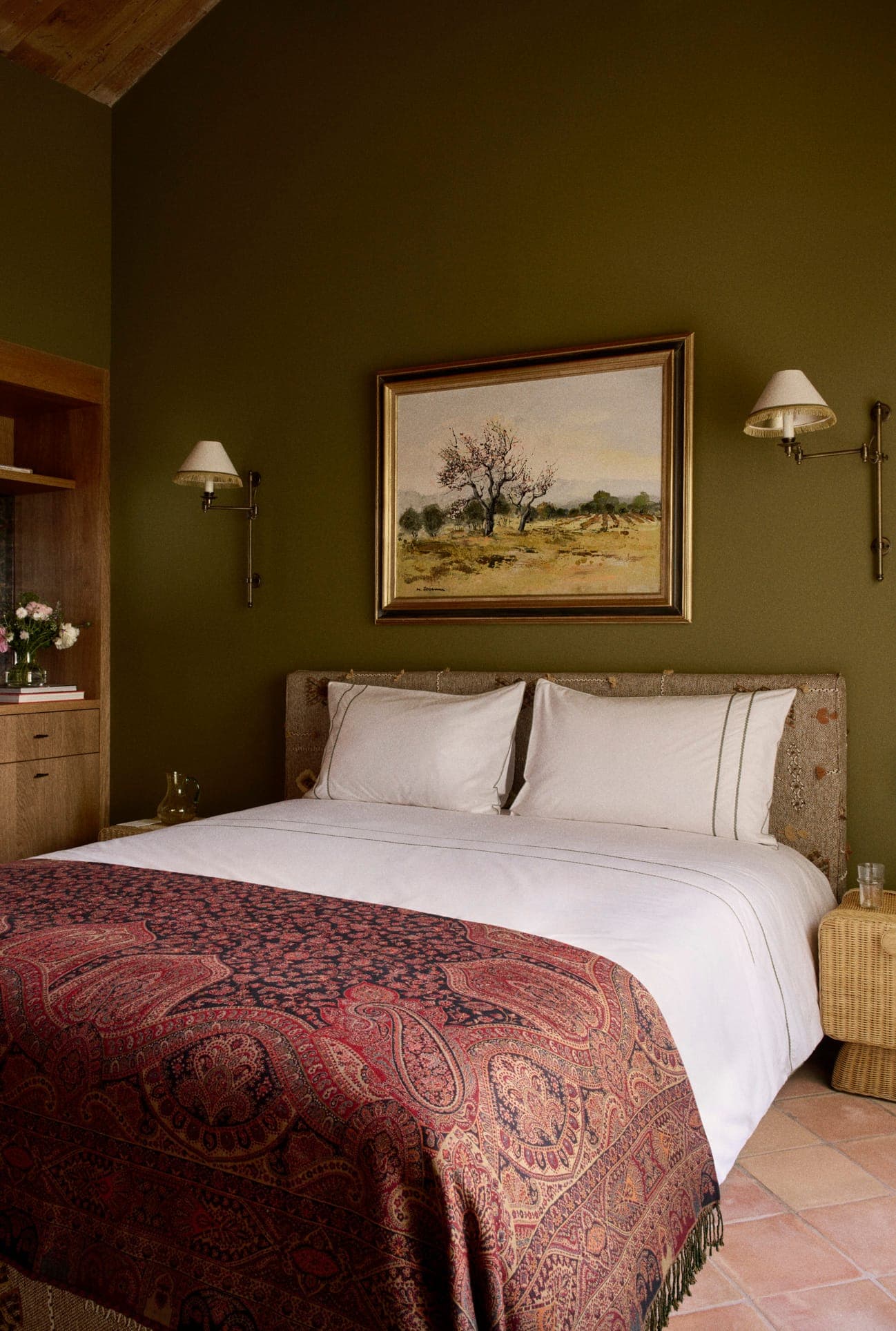 Une chambre confortable comprend un lit avec des draps blancs et une couverture à motifs. Les murs verts sont ornés d'un tableau de paysage au-dessus du lit. Des lampes murales encadrent le lit et une table de nuit en bois se trouve à côté.
