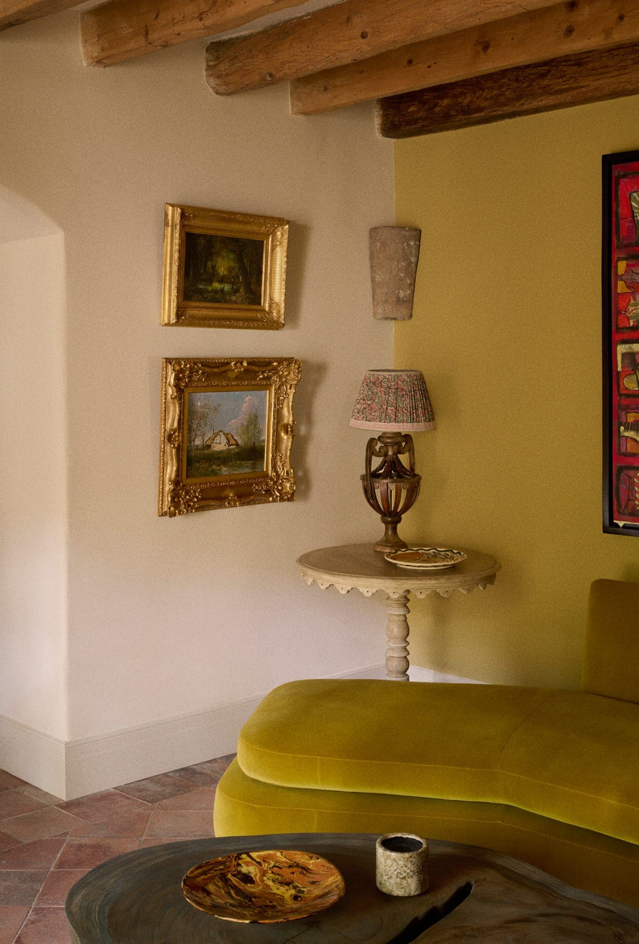Un salon chaleureux avec poutres rustiques et un canapé jaune moutarde. Deux tableaux encadrés sont accrochés sur un mur beige au-dessus d'une table d'appoint ronde avec une lampe décorative. Un grand tableau aux teintes rouges est partiellement visible à droite.