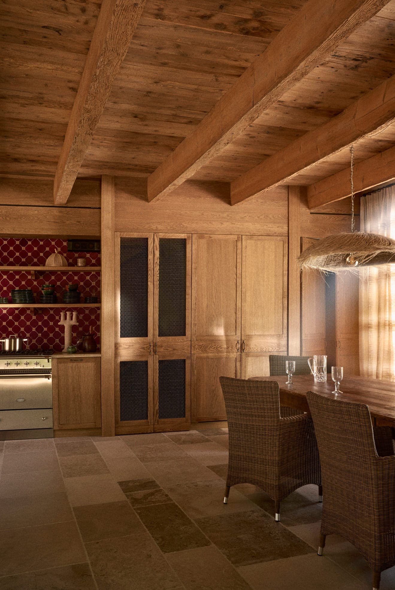 Une cuisine rustique avec poutres en bois comprend une table à manger et des chaises en osier. Elle est dotée d'un four en acier inoxydable, d'un dosseret marron et de grandes armoires en bois. La lumière chaude du soleil filtre à travers une fenêtre transparente, créant une ambiance chaleureuse.