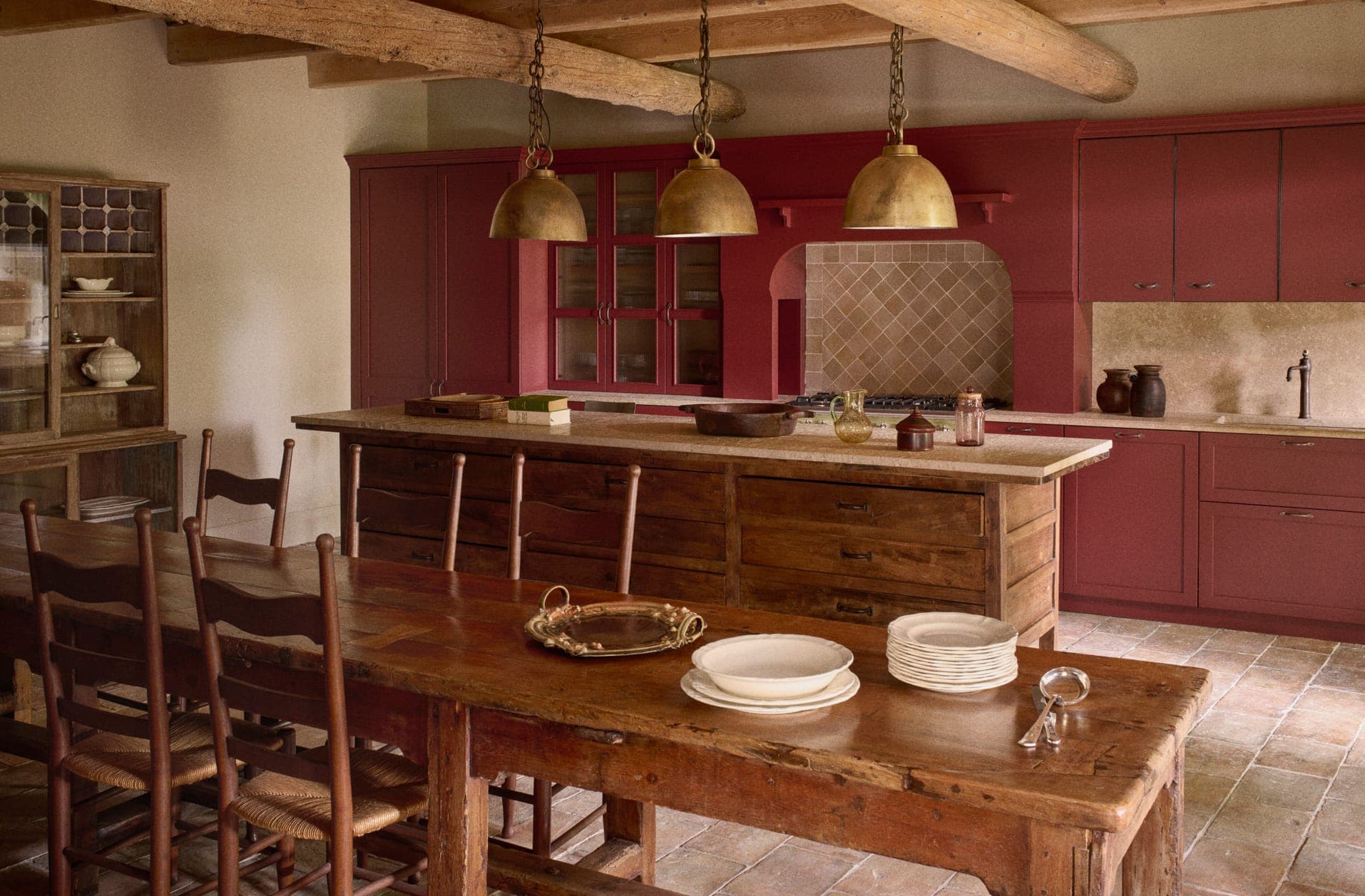 Une cuisine rustique avec des armoires rouges et un îlot en bois. Trois suspensions en laiton sont suspendues au-dessus de l'îlot. Une table à manger en bois est dressée avec des assiettes et des couverts. Des poutres en bois apparentes au plafond ajoutent à l'ambiance chaleureuse.