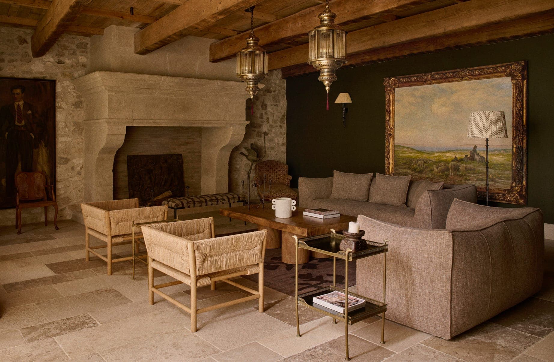 Un salon rustique avec une cheminée en pierre, deux chaises tressées, un canapé gris et une table basse en bois. La pièce est ornée de grands tableaux sur les murs, de poutres en bois au plafond et de lanternes suspendues, créant une atmosphère chaleureuse et vintage.
