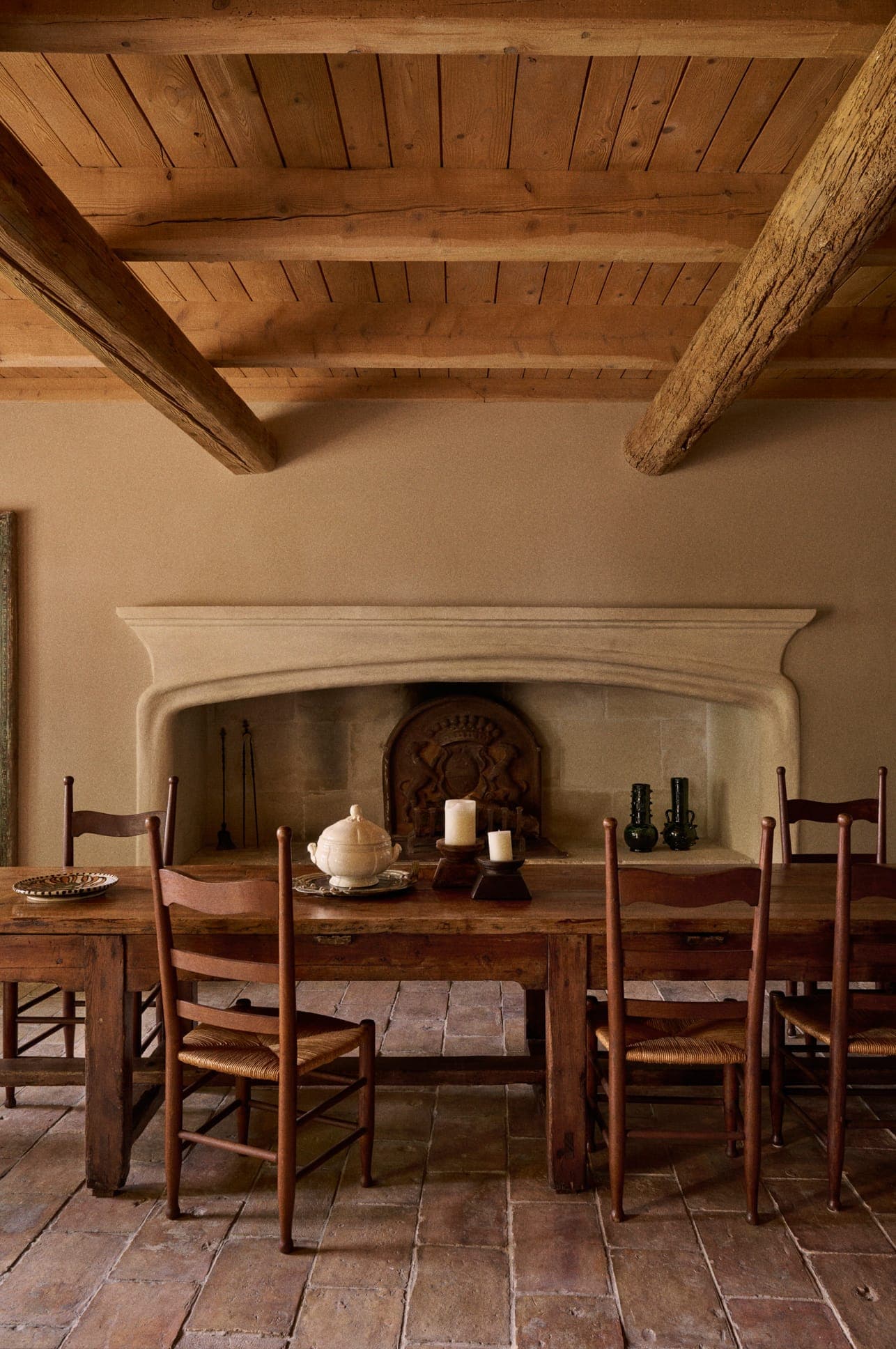 Une salle à manger rustique avec une table et des chaises en bois, avec une théière, des bougies et un set de table. L'arrière-plan présente une grande cheminée en pierre et un plafond à poutres apparentes en bois. Le sol est carrelé en terre cuite.