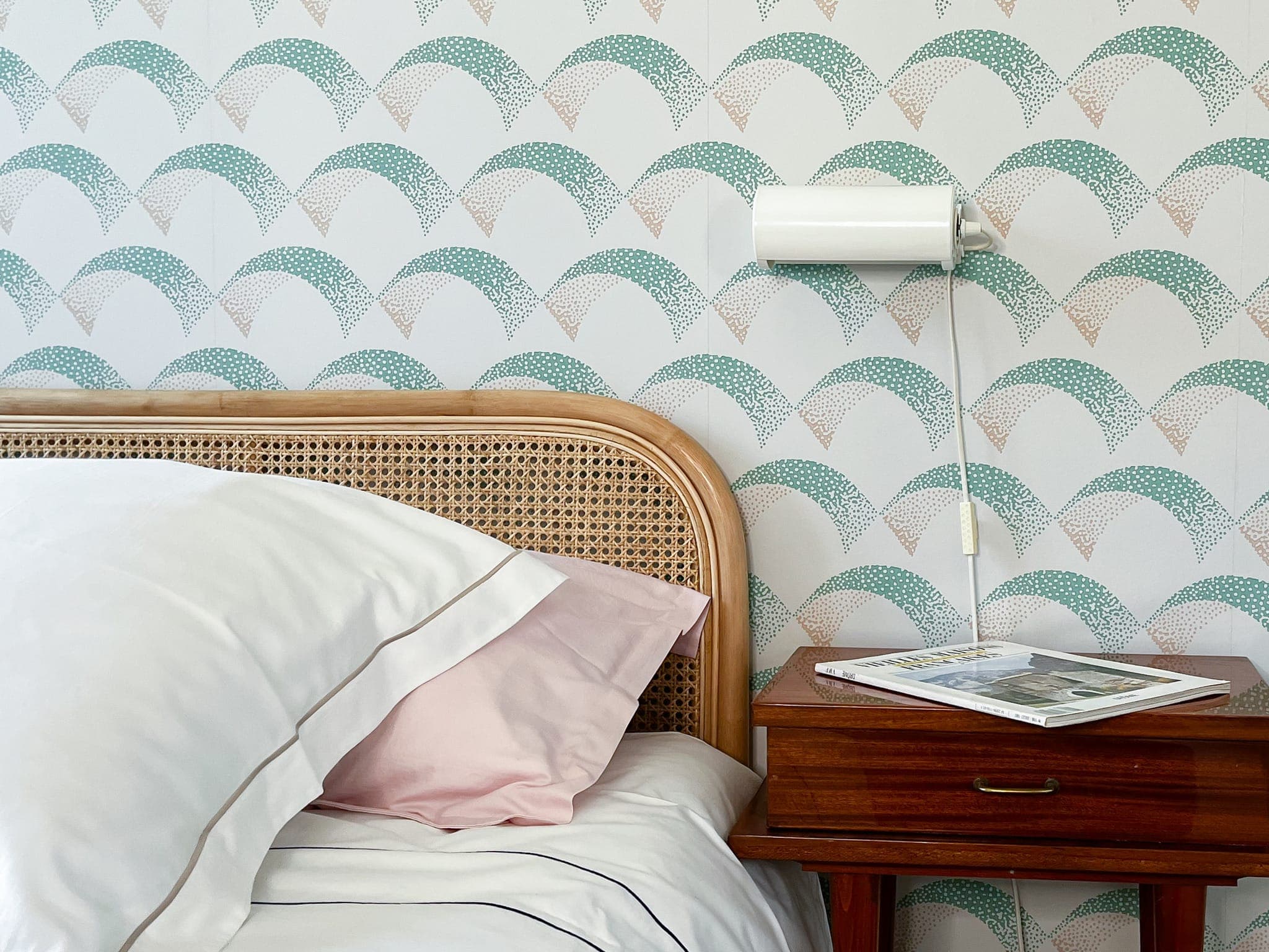 Un coin chambre est doté d'une tête de lit en rotin avec des coussins blancs et roses, d'une table d'appoint en bois avec un magazine et d'une applique murale blanche. Le mur est orné d'un motif en arcs bleu sarcelle, pêche et blanc.