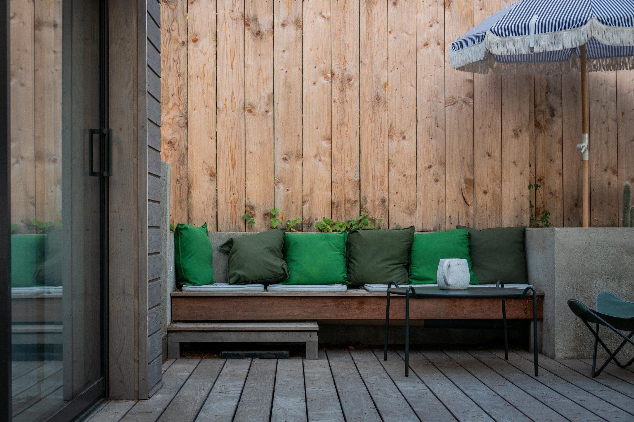 A cozy outdoor terrace with a wooden bench lined with green cushions. A small white vase sits on a coffee table in front. The space is enclosed by a wooden fence, and a blue and white striped umbrella provides shade.