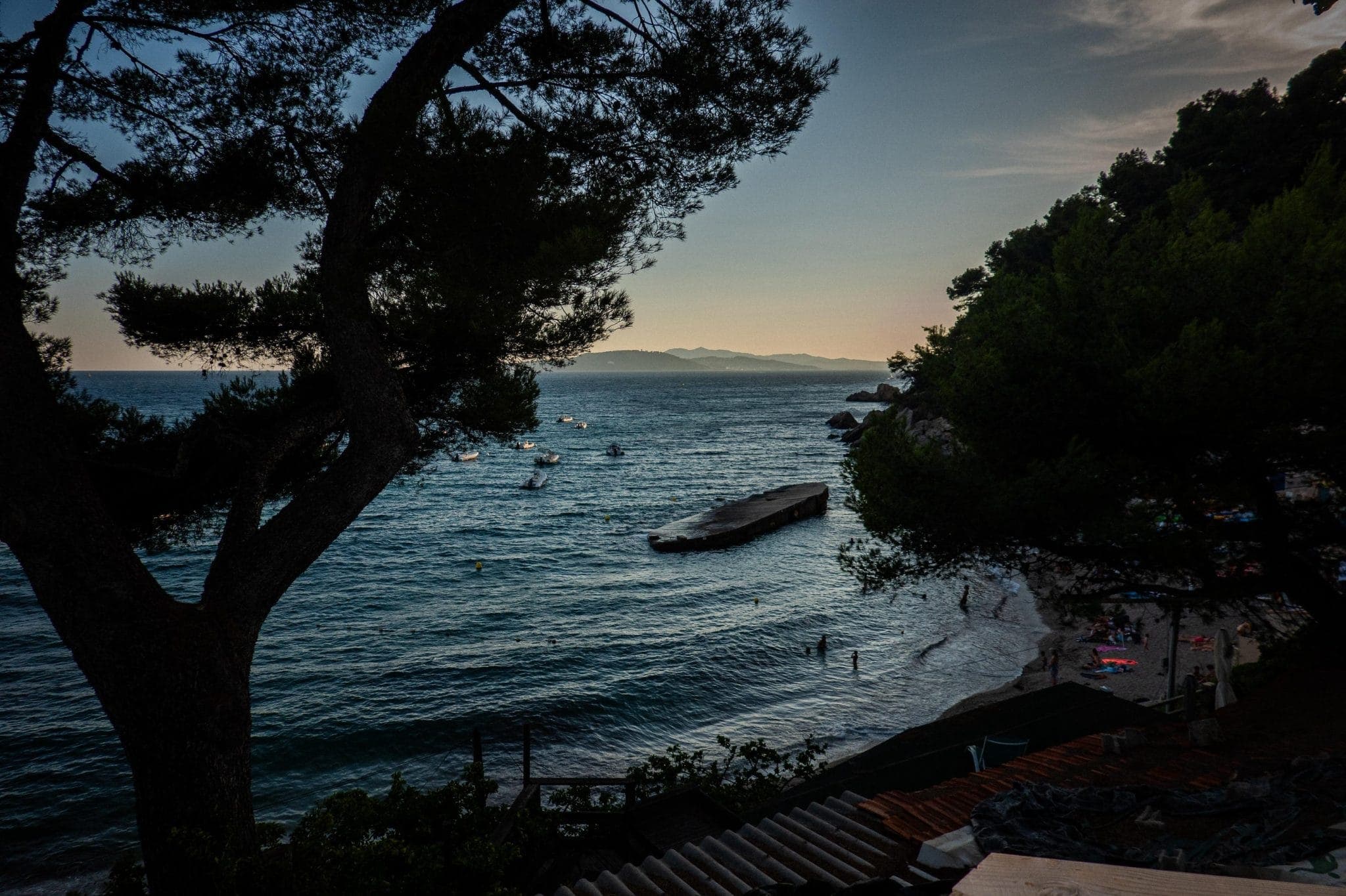 A serene view of the coast at dusk, with gentle waves lapping at the shore. Silhouetted trees frame the scene, and a few boats float in the distance. The sky transitions from warm hues to darker tones as night approaches.