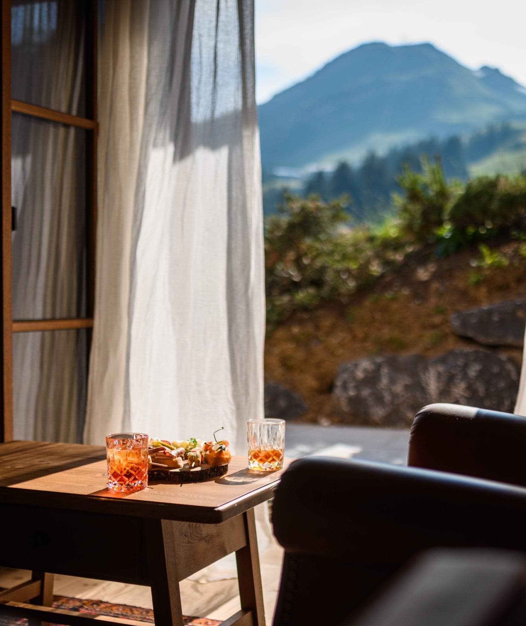 Une scène d'intérieur chaleureuse avec une petite table en bois sur laquelle sont posés deux boissons et un plateau de collations, près d'une porte ouverte. La lumière du soleil entre à flots et une montagne verdoyante est visible en arrière-plan, créant une atmosphère sereine et accueillante.
