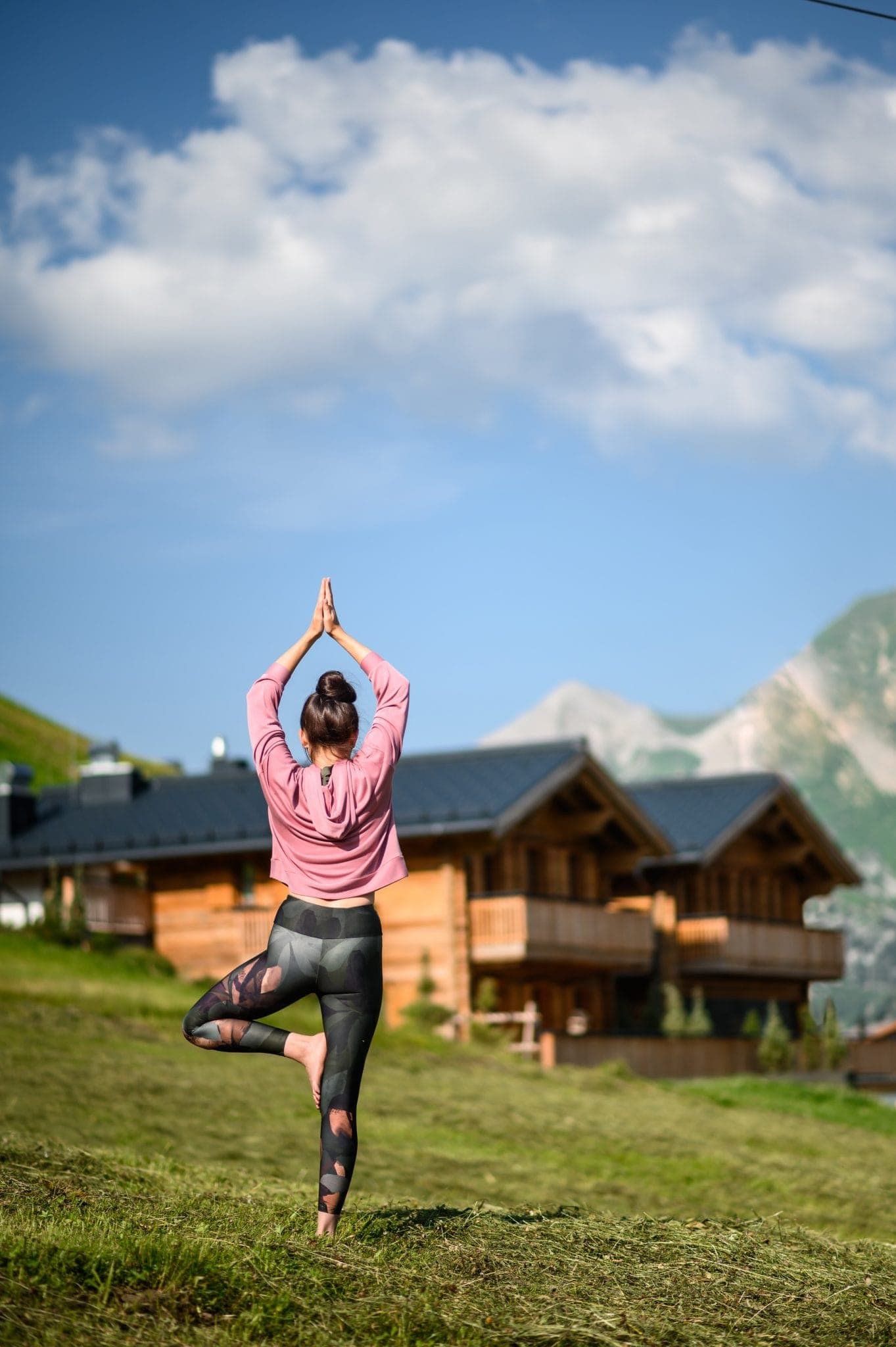 Une personne pratique le yoga dans la posture de l'arbre sur une colline herbeuse. Elle porte un haut rose et des leggings à motifs. En arrière-plan, il y a des cabanes en bois et des montagnes sous un ciel bleu parsemé de nuages.
