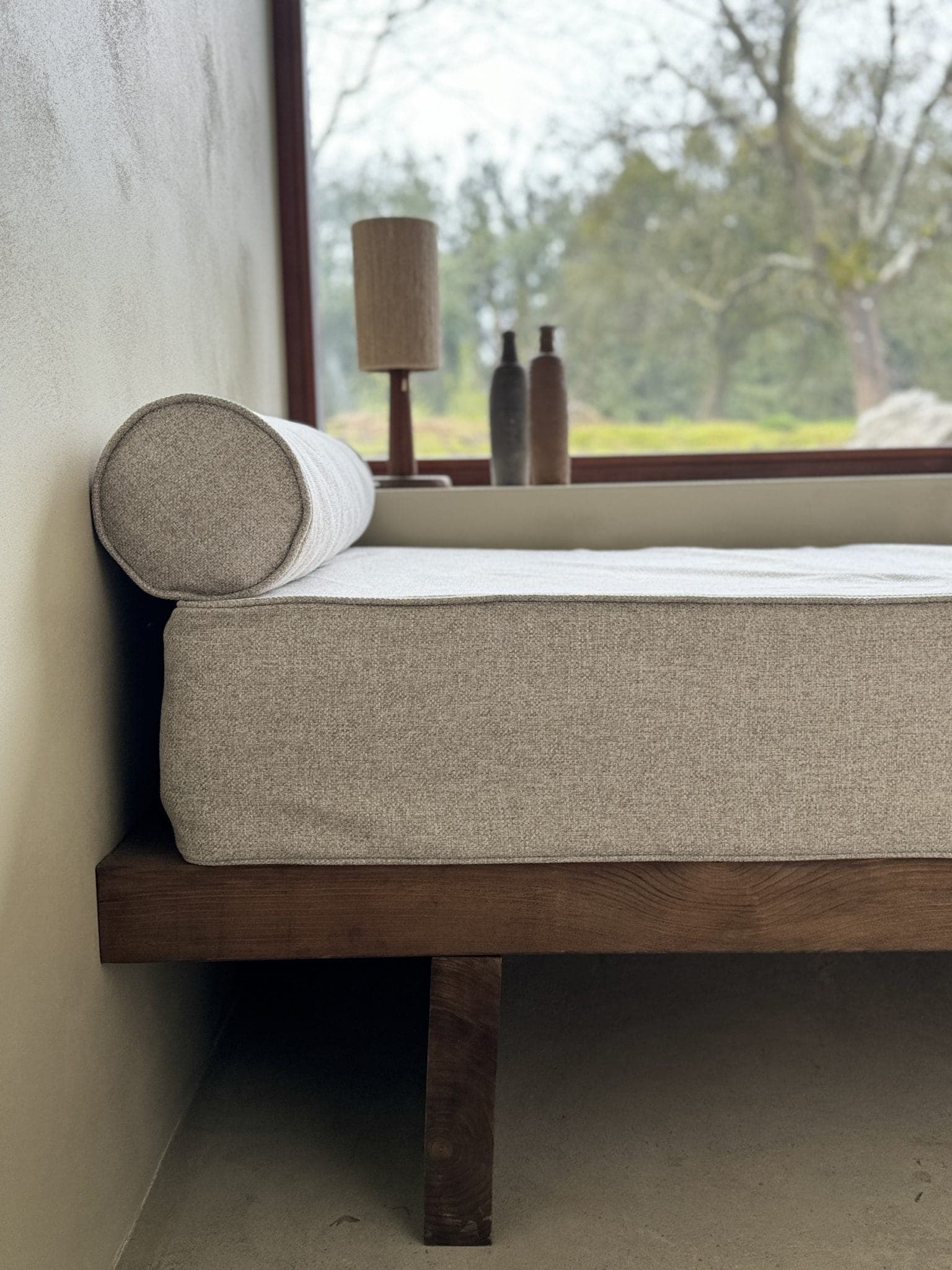 Chambre minimaliste avec un cadre de lit en bois et un matelas beige. Un oreiller en rouleau accentue le lit. Une lampe et deux vases sont posés sur le rebord de la fenêtre, avec des arbres visibles à l'extérieur. La lumière naturelle illumine les tons neutres.
