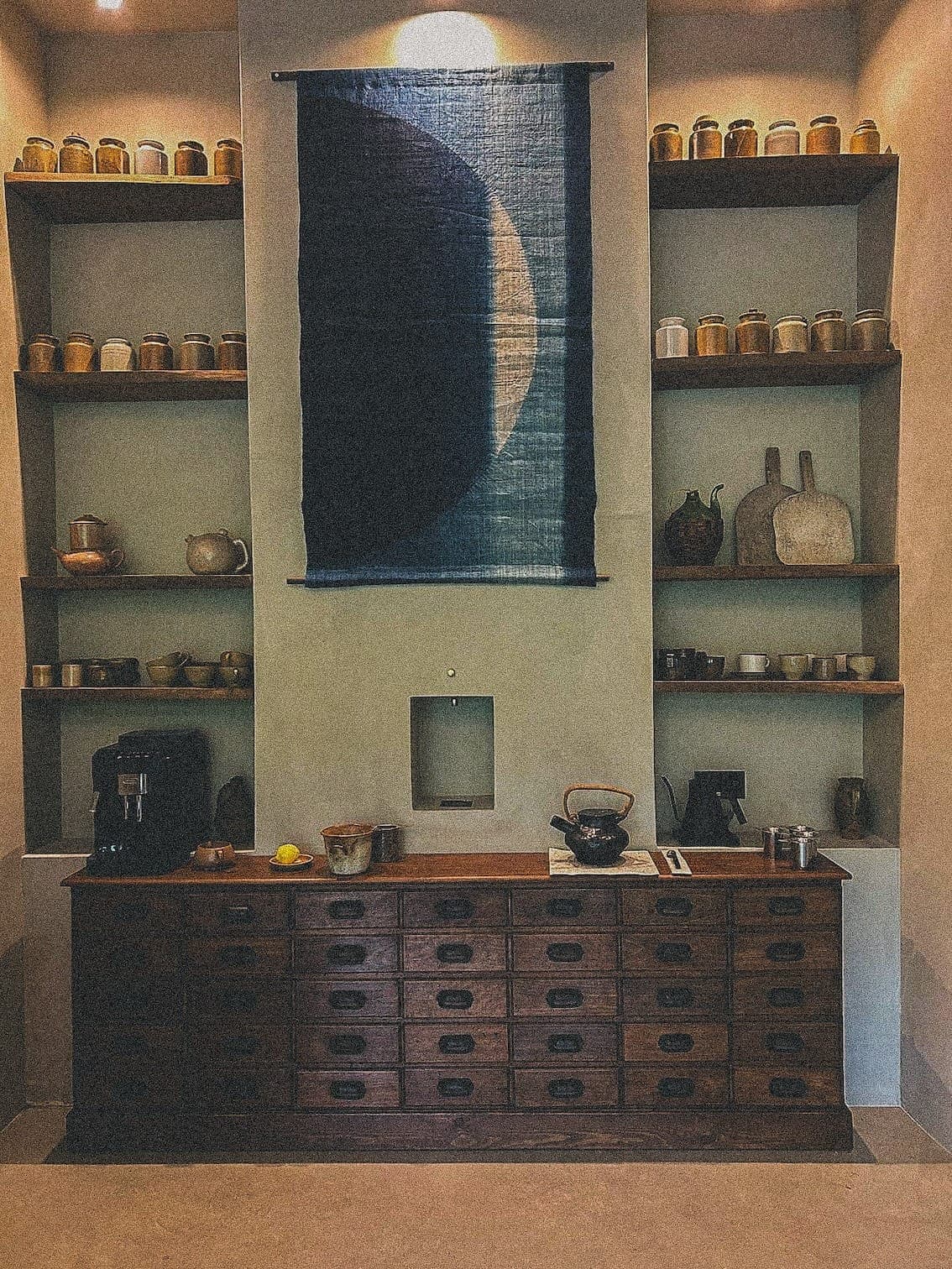 Une scène de cuisine rustique avec un meuble en bois, décoré de poteries et d'ustensiles de cuisine. Un grand textile indigo avec un motif de croissant de lune est suspendu au-dessus. Des étagères de chaque côté contiennent des bocaux et d'autres poteries, créant une ambiance chaleureuse et vintage.
