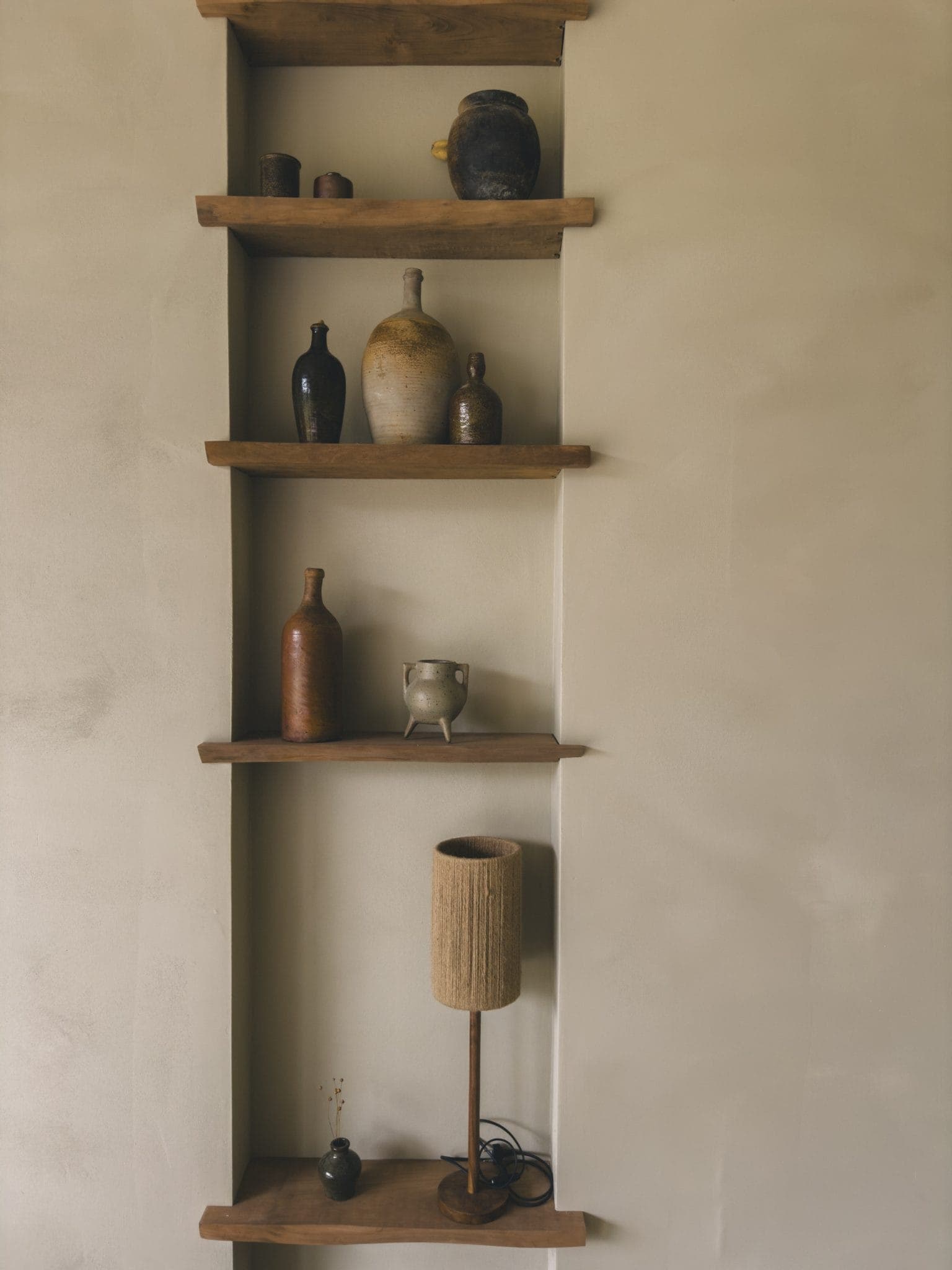 Étagères murales en bois présentant des poteries rustiques, des vases et une petite lampe contre un mur de couleur claire. Les étagères ont une esthétique minimaliste et terreuse.