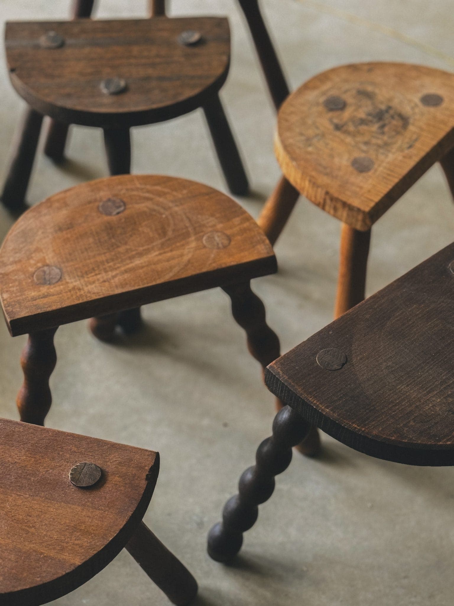 Une collection de cinq tabourets en bois avec des sièges ronds et en demi-lune disposés sur un sol en béton. Les tabourets ont une apparence rustique avec différentes nuances de brun et présentent des assemblages à chevilles simples et ronds.