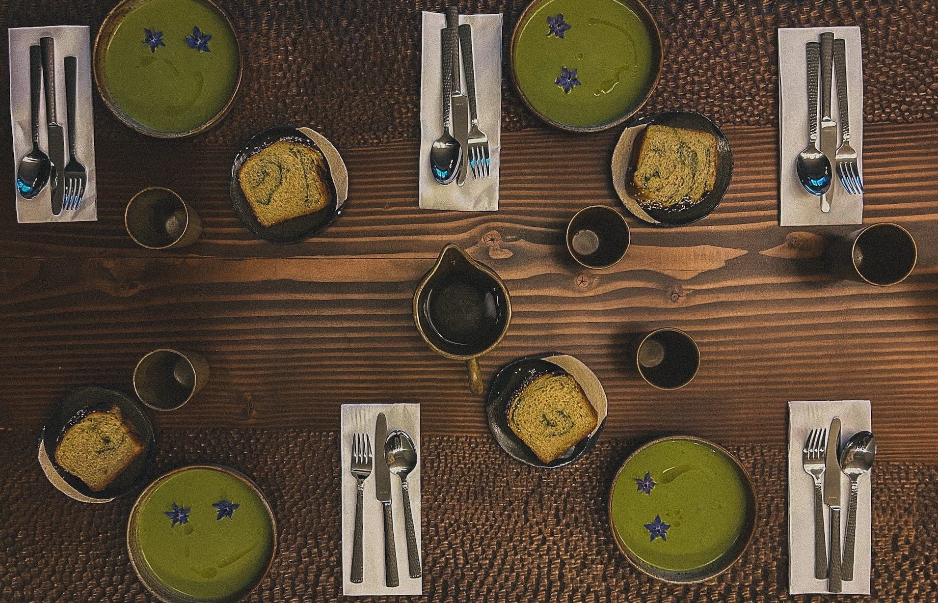 Une table en bois dressée pour six personnes avec des bols de soupe verte et des tranches de pain sur des assiettes sombres. Chaque service comprend une fourchette, un couteau, une cuillère et une serviette. De petites fleurs violettes décorent la soupe, et il y a trois grandes tasses et une cruche noire au centre.