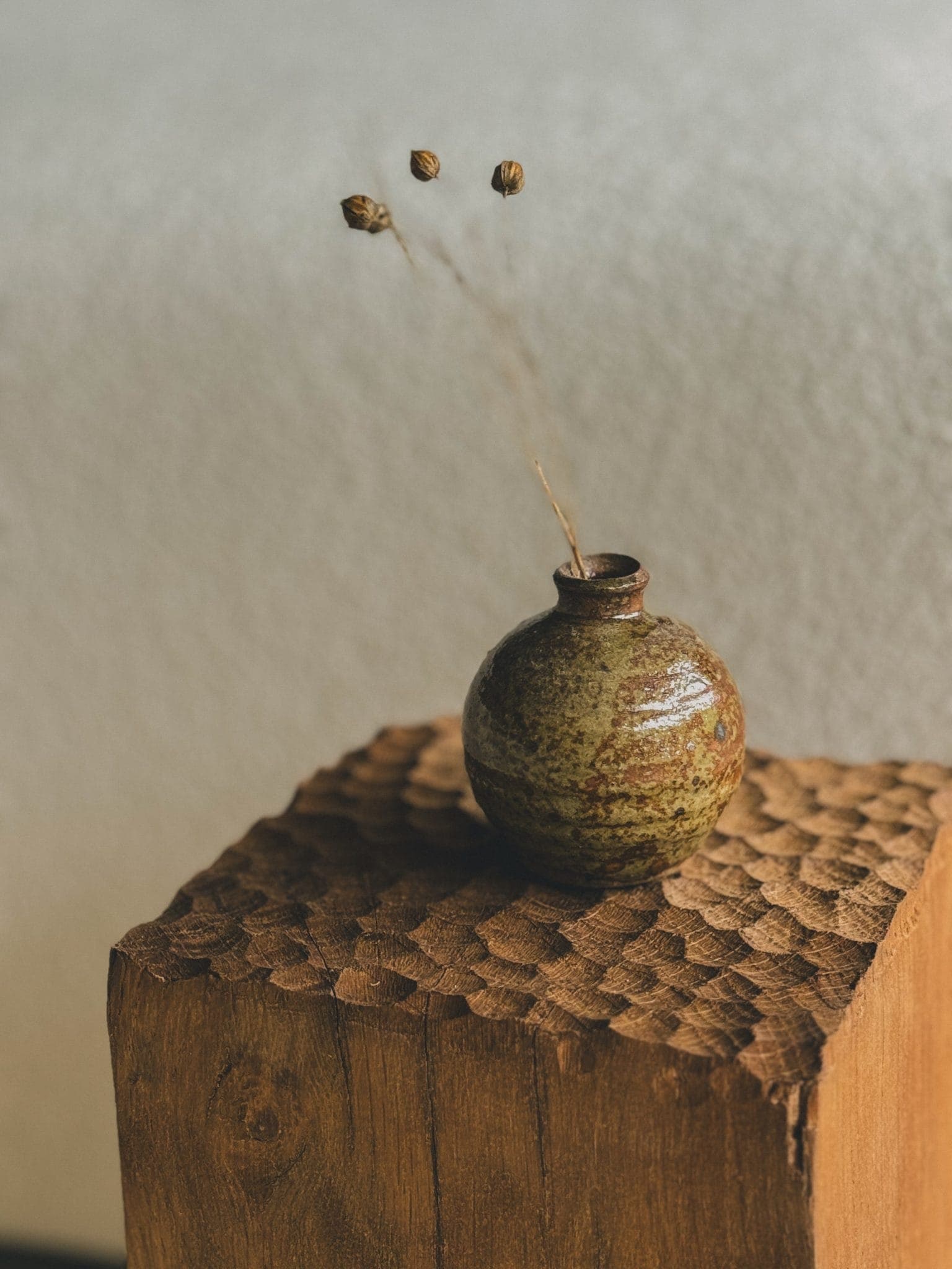 A small round ceramic vase with a textured, earthy glaze rests on a carved wooden block. Dried stems with buds extend from the vase. The background is softly blurred, creating a serene atmosphere.