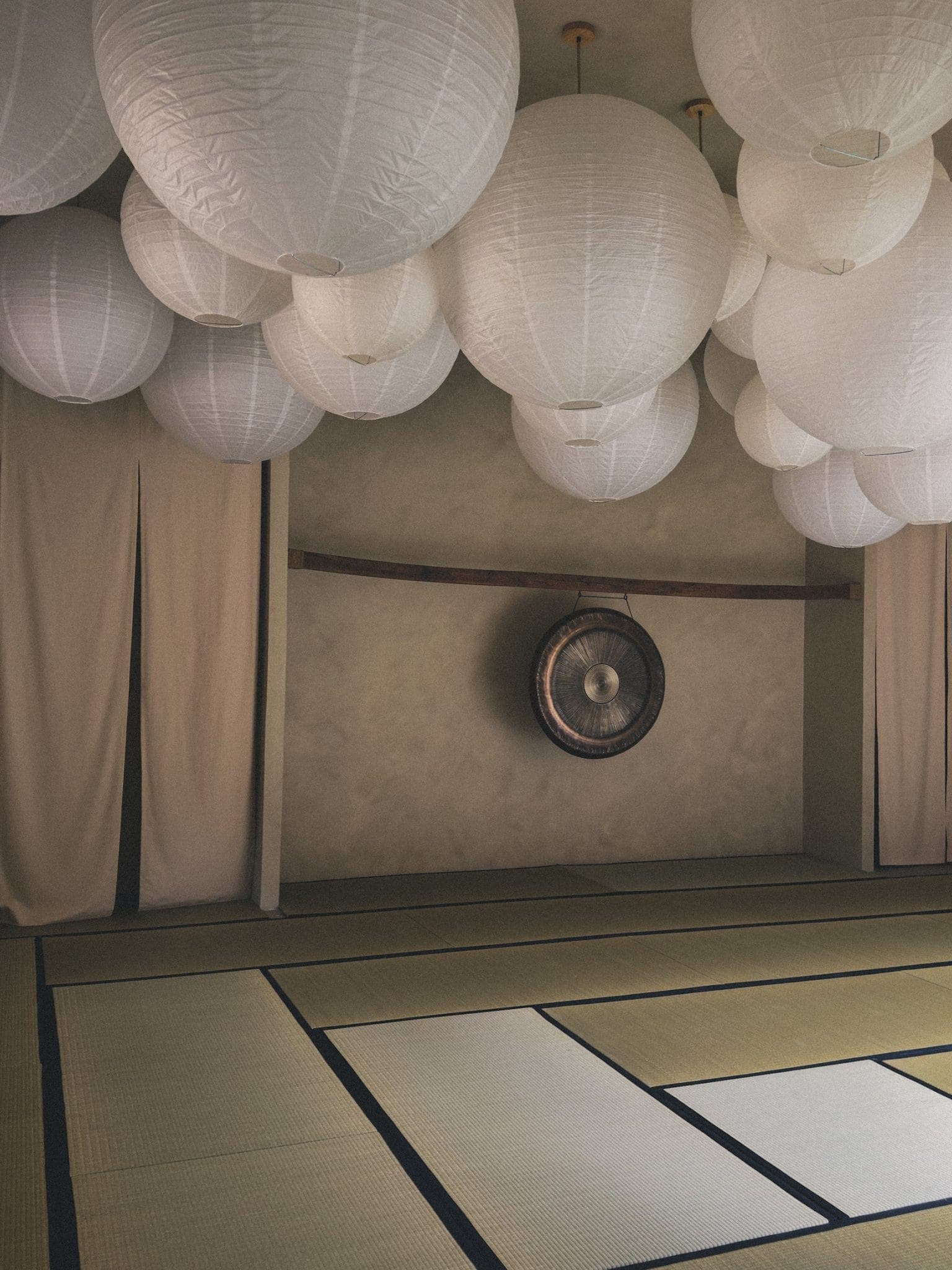 Une pièce sereine avec de grandes lanternes en papier suspendues au plafond, un gong sur le mur et un sol recouvert de tatamis. Les murs sont de couleur neutre et des rideaux drapés pendent sur les côtés.