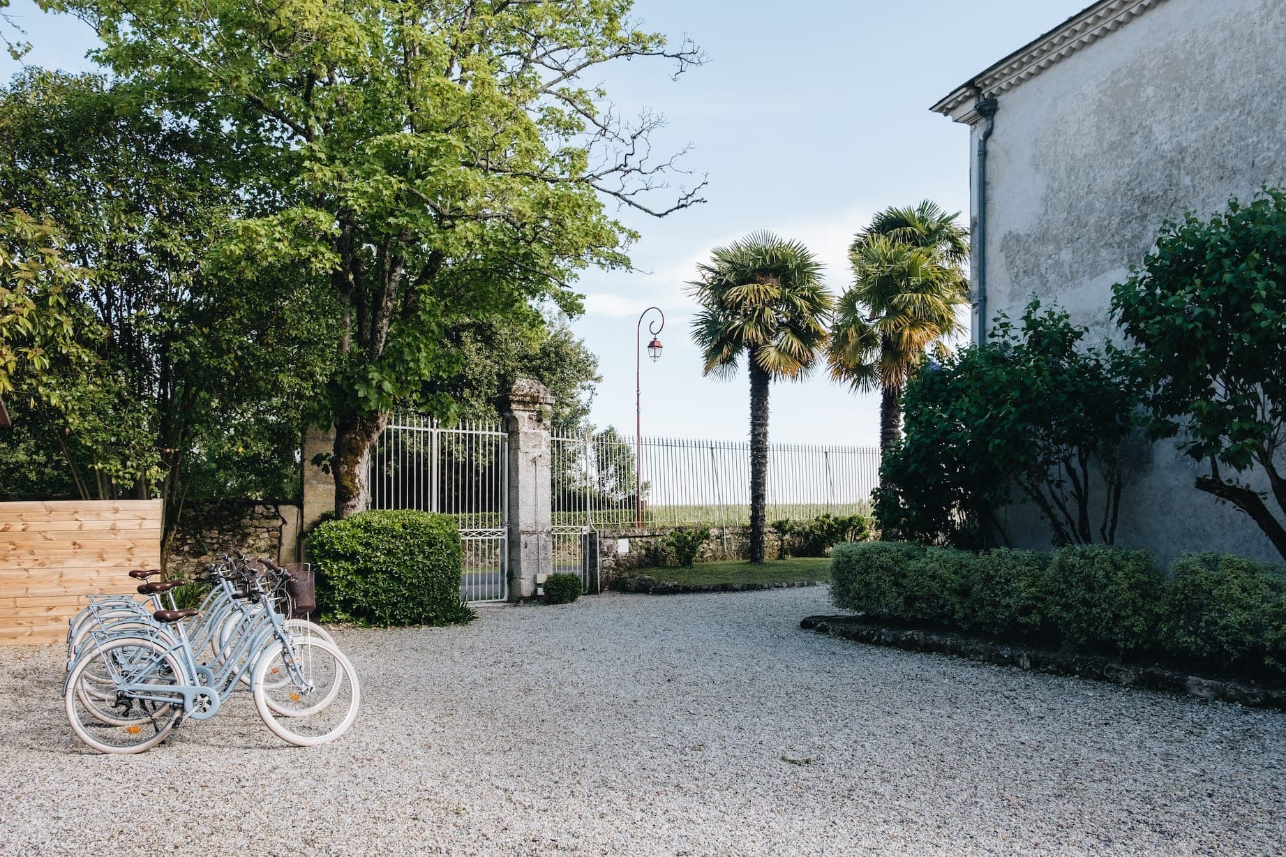 Une scène extérieure sereine avec une allée de gravier menant à un portail ouvert en fer forgé. Trois vélos de style vintage reposent sur le gravier. Une végétation luxuriante, comprenant des arbres et des buissons, entoure la zone, et un haut mur enferme partiellement l'espace.