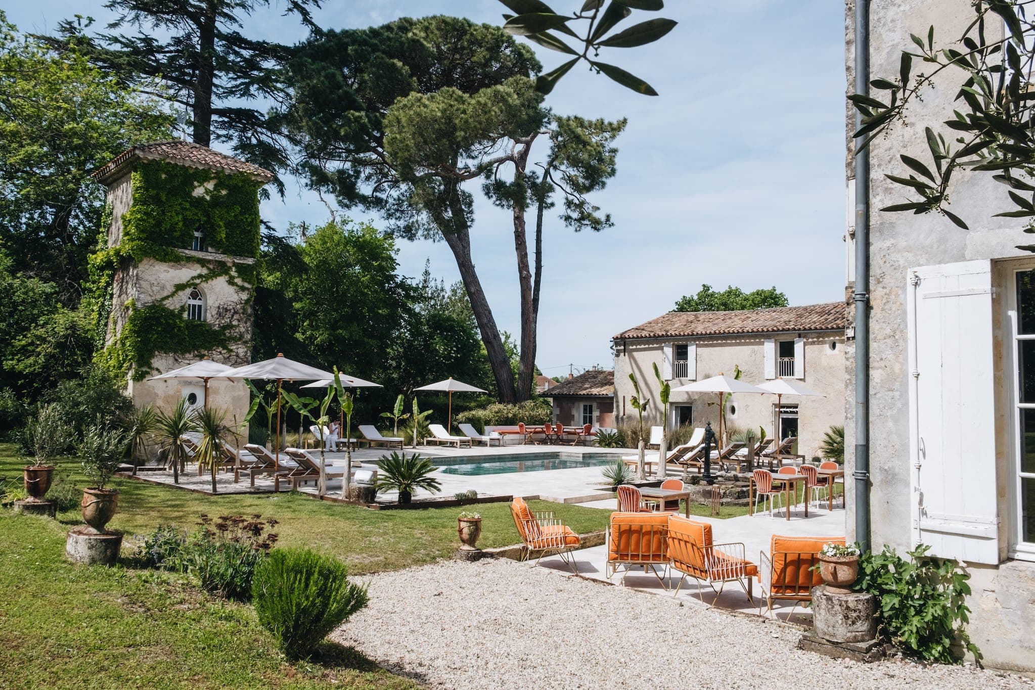 Une charmante cour intérieure avec des bâtiments anciens entourés d'arbres verts. Au centre, se trouve une piscine avec des chaises longues et des parasols. Des chaises orange sont disposées dans un espace gazonné, créant une atmosphère sereine et accueillante.