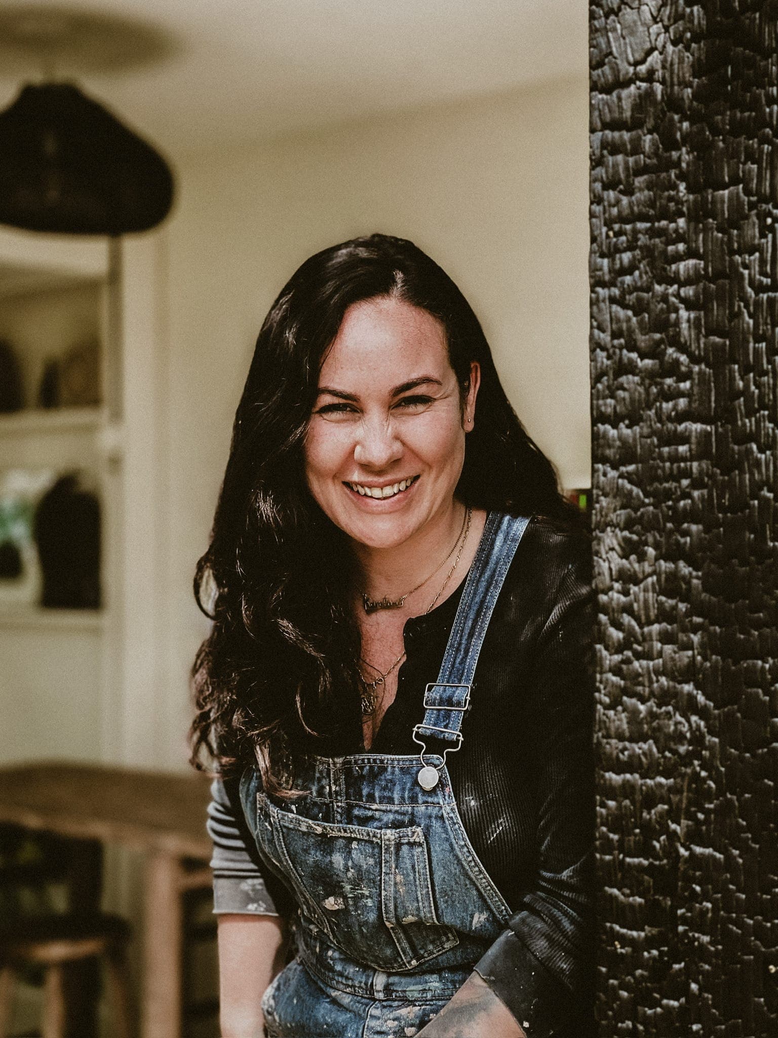 Karolänn stands outdoors, her long dark hair flowing as she smiles warmly against a blackened wooden post. Dressed in a black top and denim overalls, the scene is completed by a wooden table and light-colored walls, setting the perfect backdrop for this moment of contentment.
