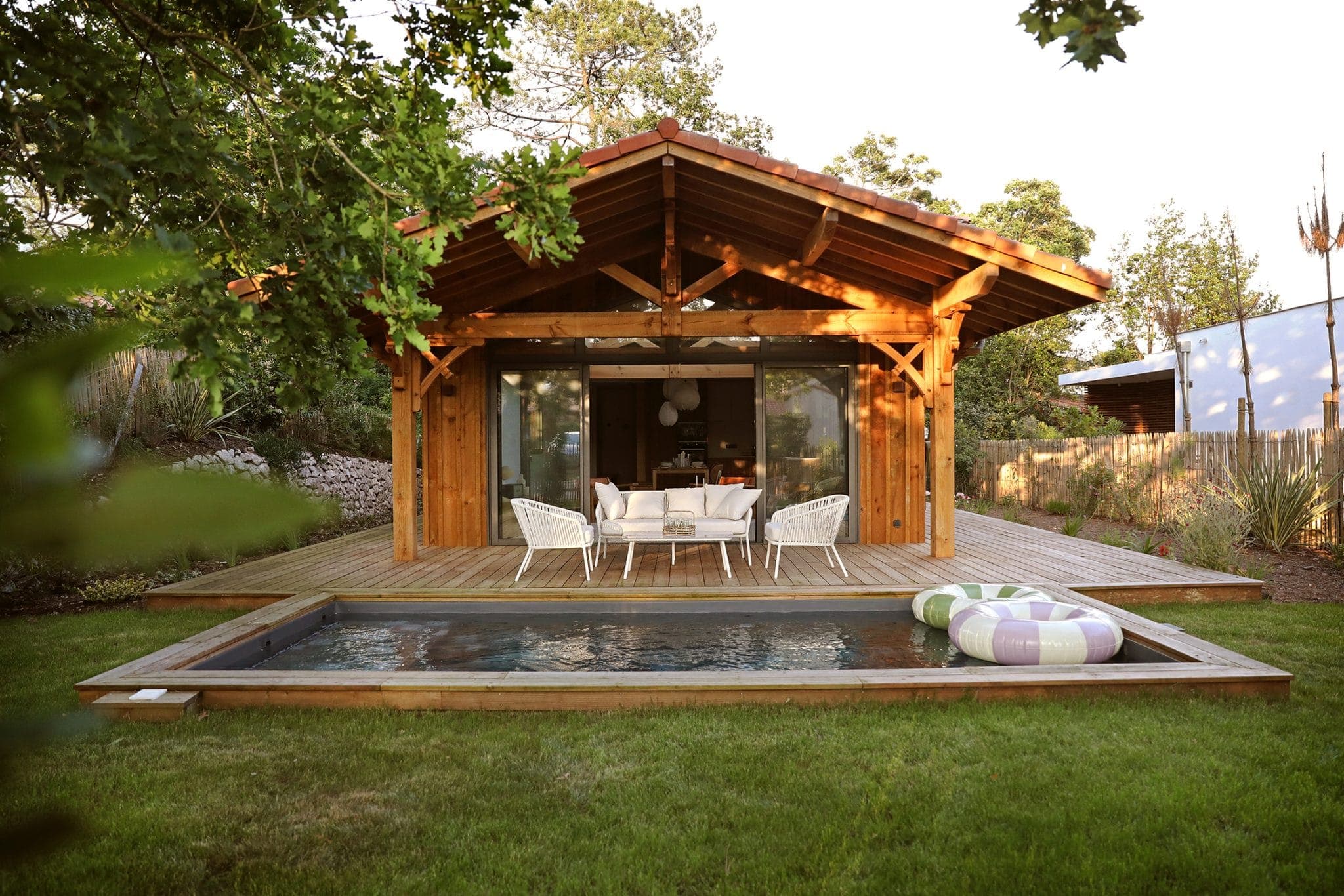 A cozy wooden cabin with a large deck sits by a small rectangular pool. The cabin features a covered outdoor dining area with white chairs. Surrounded by grass and trees, the scene is peaceful and inviting.