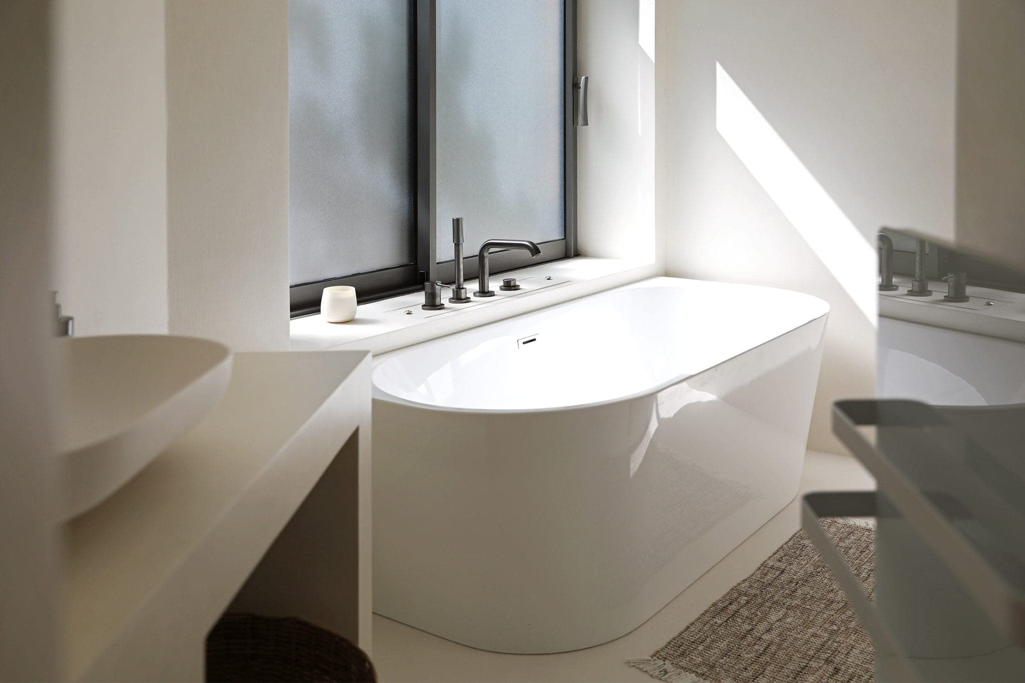 A modern bathroom features an elegant white bathtub set next to a large frosted window. The bath has a minimalist faucet and a small candle beside it, with soft natural light flooding in, creating a serene, welcoming atmosphere.