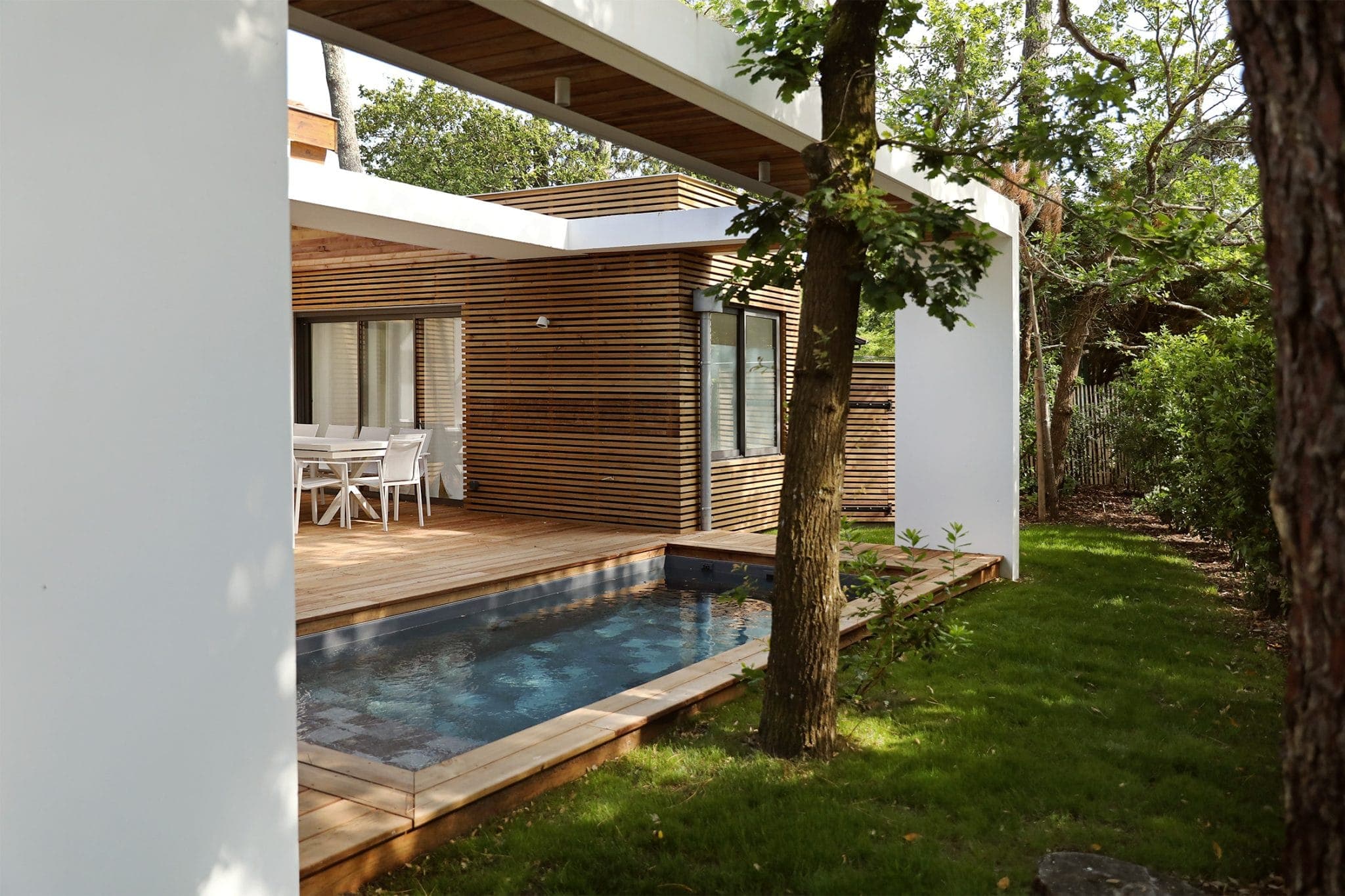 The exterior of a modern house features horizontal wooden slats and large glass doors. In the foreground is a small rectangular pool surrounded by a wooden deck, with a tree and lush grass nearby. A white outdoor dining set is visible under the roof.