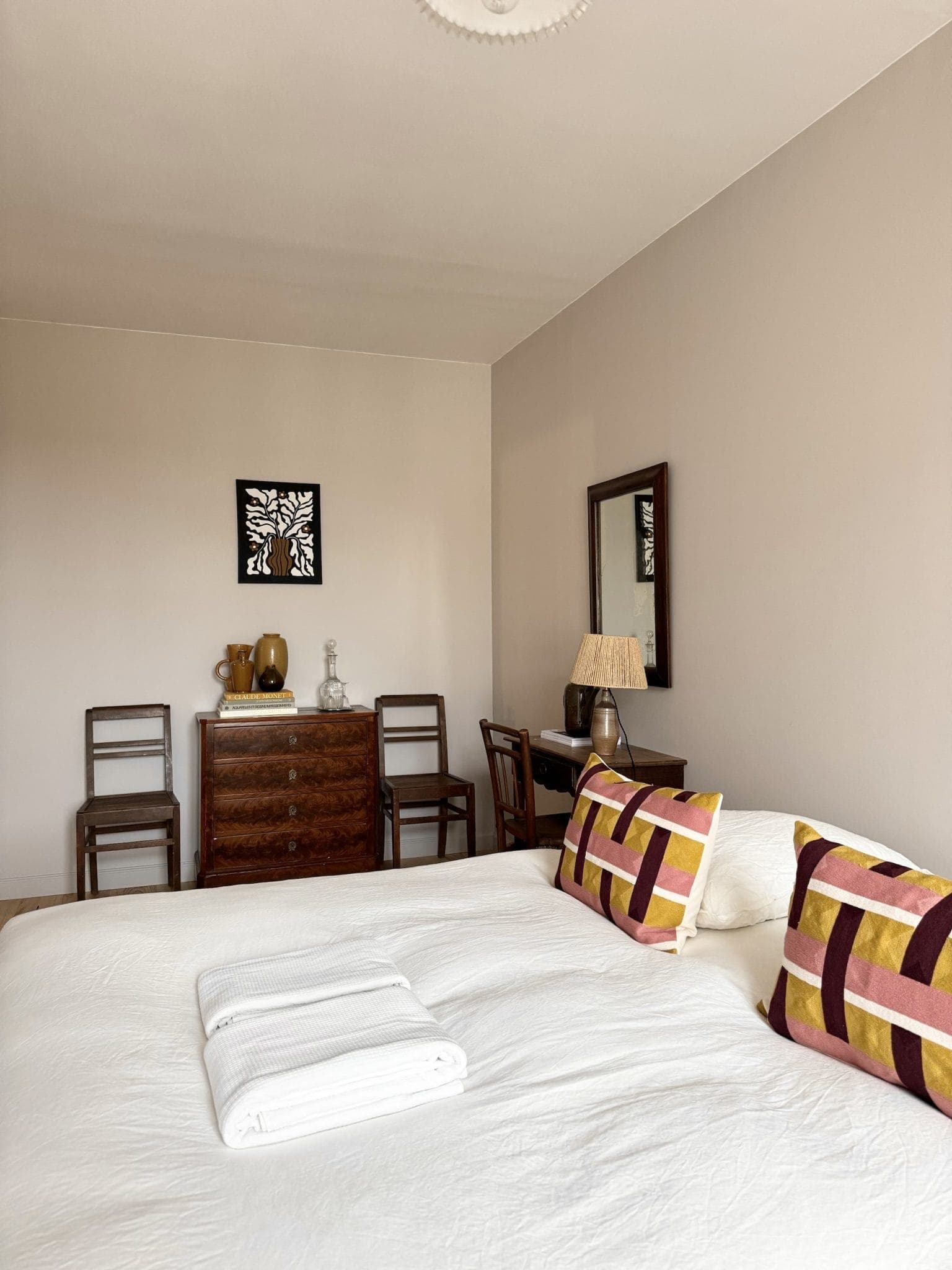 Une chambre minimaliste avec un lit bien fait avec deux oreillers géométriques et une serviette pliée. La chambre comprend une commode en bois, deux chaises, une lampe et des œuvres d'art murales. Les tons neutres créent une atmosphère calme.