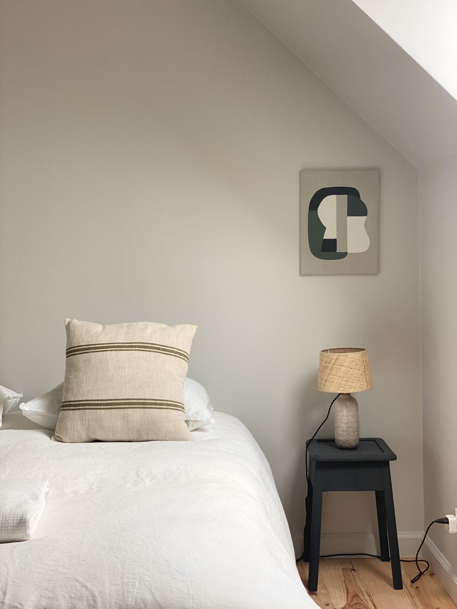 Une chambre minimaliste avec un lit blanc et un coussin à rayures beiges, à côté d'une table d'appoint noire avec une lampe texturée. Une œuvre d'art abstraite est accrochée au mur blanc en angle. Le parquet en bois clair renforce l'impression d'espace de la pièce.