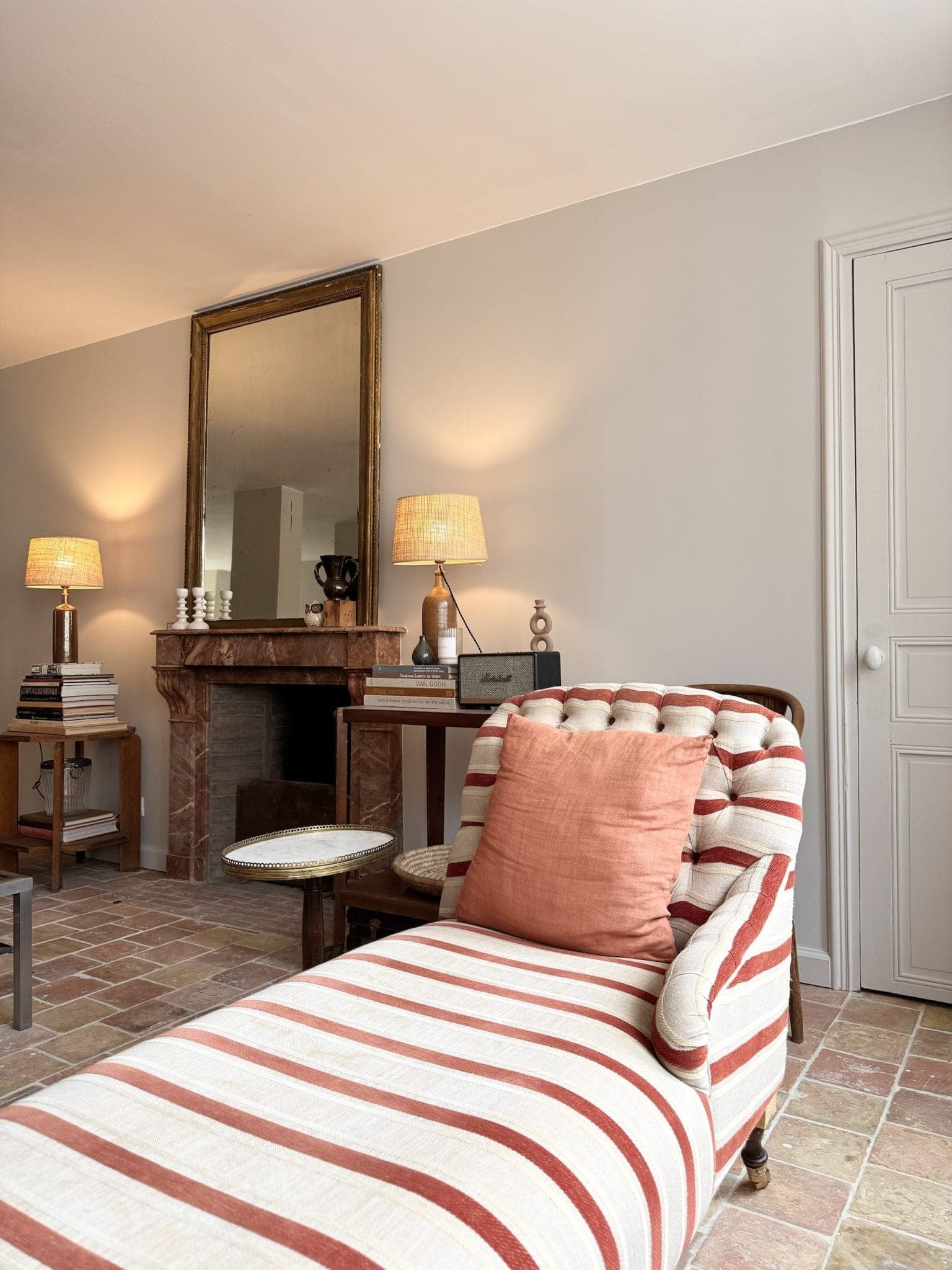 Un salon chaleureux avec une chaise longue rayée et un coussin orange au premier plan. Derrière, une cheminée en bois surmontée d'un grand miroir, flanquée de deux lampes, crée une ambiance chaleureuse. Le sol est carrelé dans des tons terreux.
