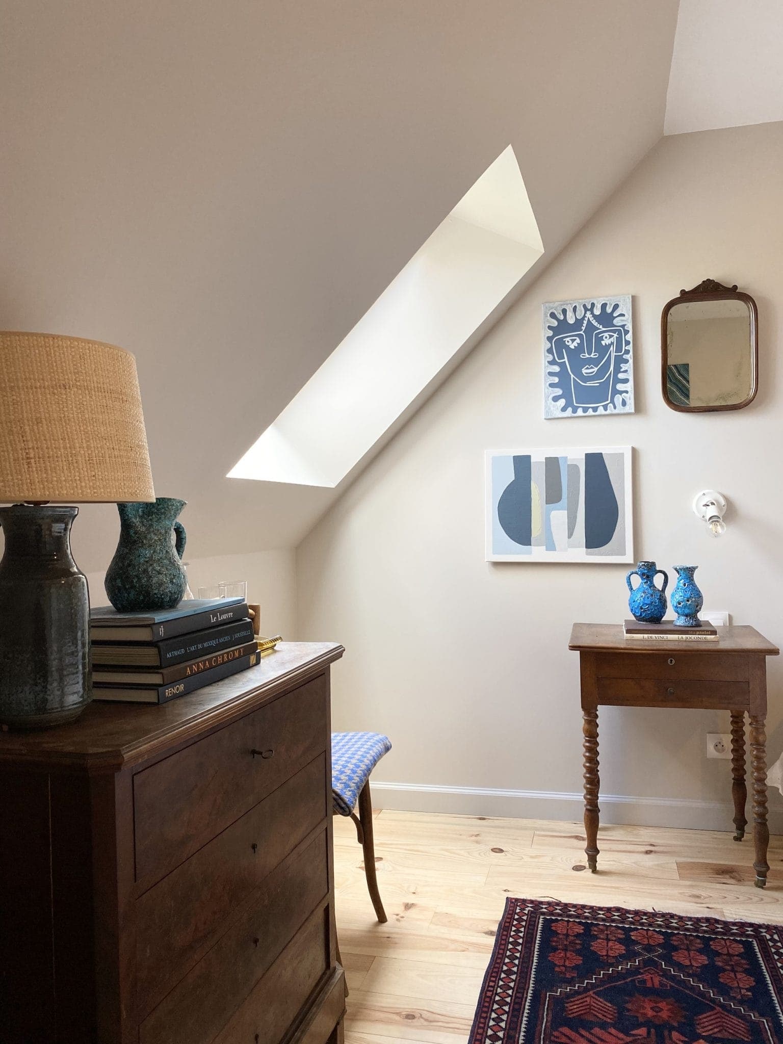 Une pièce confortable est dotée d'un plafond incliné et d'une lucarne. Une commode en bois abrite des livres, un vase bleu et une lampe. À proximité, une petite table supporte des pichets bleus assortis. Deux œuvres d'art abstraites sont accrochées au mur au-dessus et un miroir décoratif est également présent.