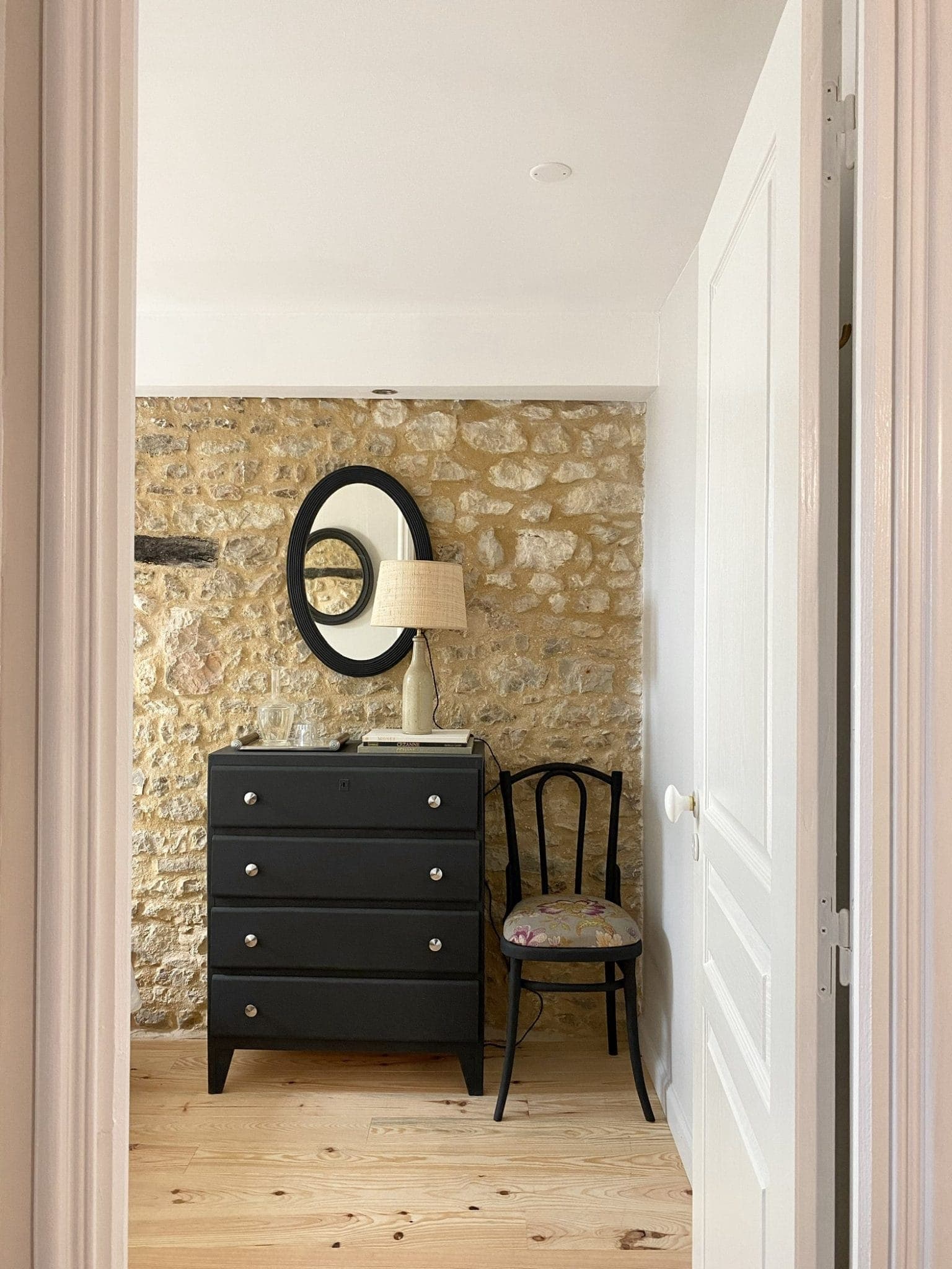 Une pièce chaleureuse avec un mur en pierre rustique avec une commode noire avec des boutons argentés, un miroir ovale au-dessus et une lampe au-dessus. Une chaise en bois avec un coussin fleuri est posée à côté de la commode sur un plancher en bois clair.