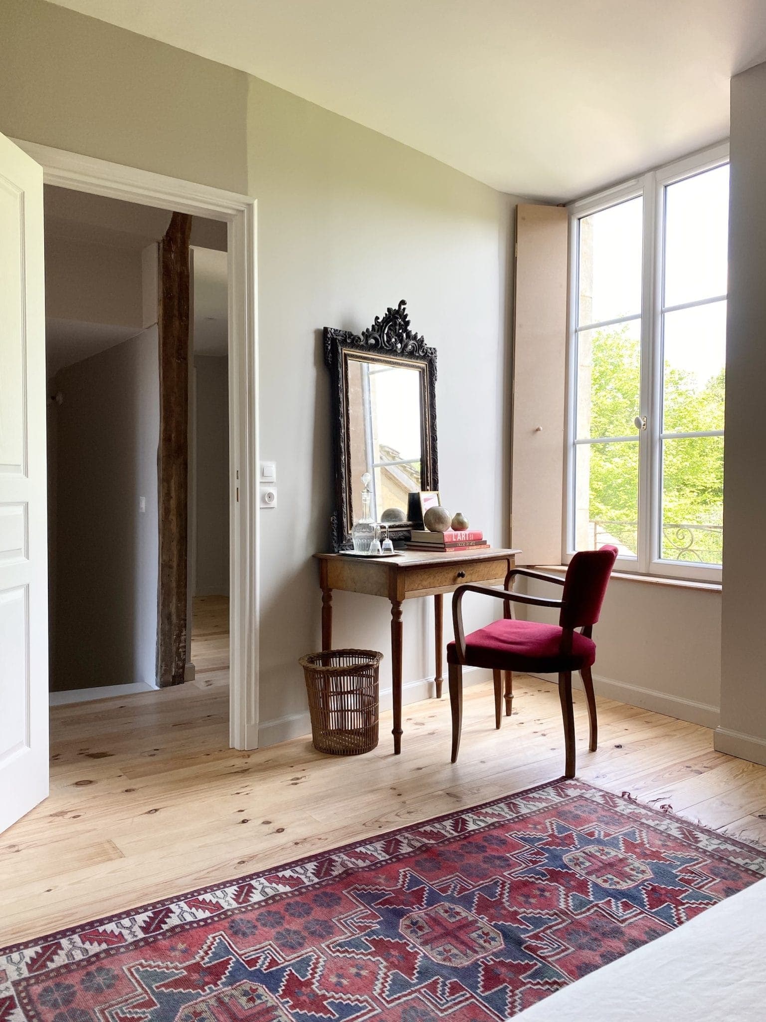 Coin chambre lumineux avec un bureau en bois et un miroir décoré. Une chaise rouge rembourrée se trouve à côté du bureau, sur lequel sont posés des articles de toilette. De grandes fenêtres laissent entrer la lumière du soleil et un tapis à motifs recouvre le sol. Porte ouverte et poutre apparente en vue.
