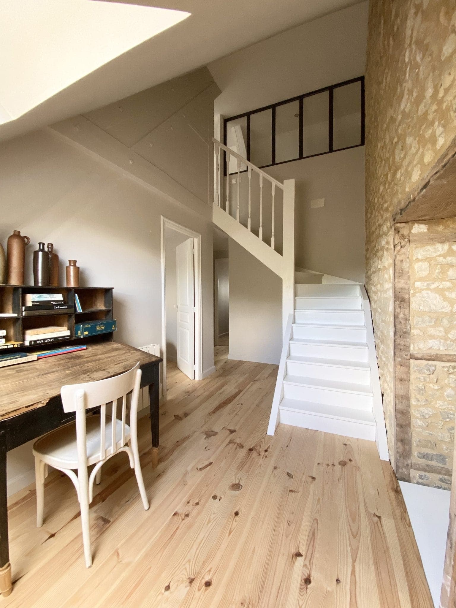 Un intérieur cosy avec un bureau et une chaise en bois sur un sol en pin clair. Un escalier aux marches blanches mène à une mezzanine avec une cloison en verre à cadre noir. Les murs sont beiges, avec un mur en pierre apparente. Des étagères accueillent des objets de décoration.