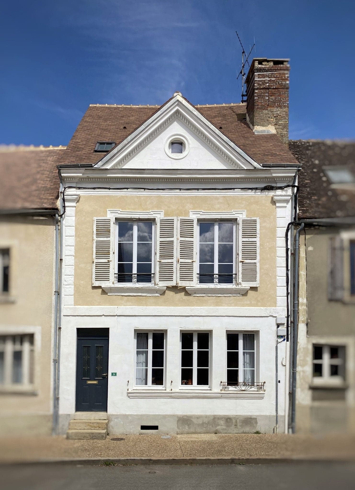 Une charmante maison à deux étages avec des murs beiges et blancs, des volets rustiques aux fenêtres supérieures et une porte bleu foncé. Le toit est doté d'une cheminée et il y a une petite fenêtre dans le pignon triangulaire. C'est une journée ensoleillée avec un ciel bleu clair.