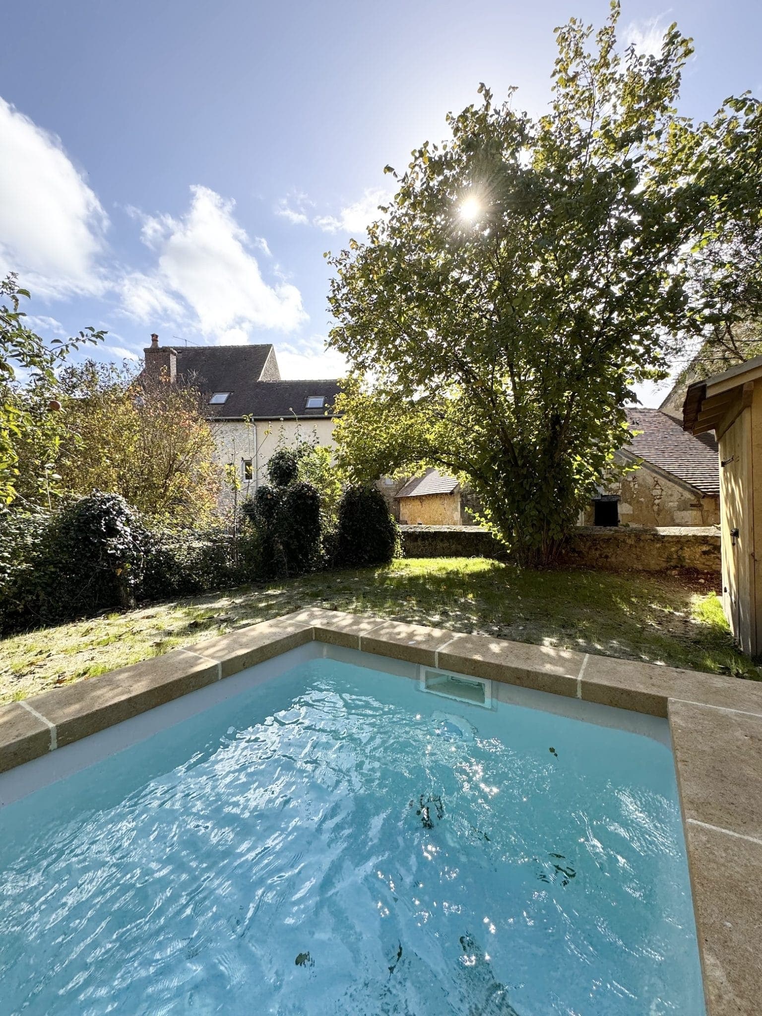 Petite piscine ensoleillée à côté d'une maison en pierre au toit en pente. Un grand arbre offre de l'ombre dans la cour gazonnée. Le ciel est clair avec quelques nuages et la lumière du soleil filtre à travers les feuilles.