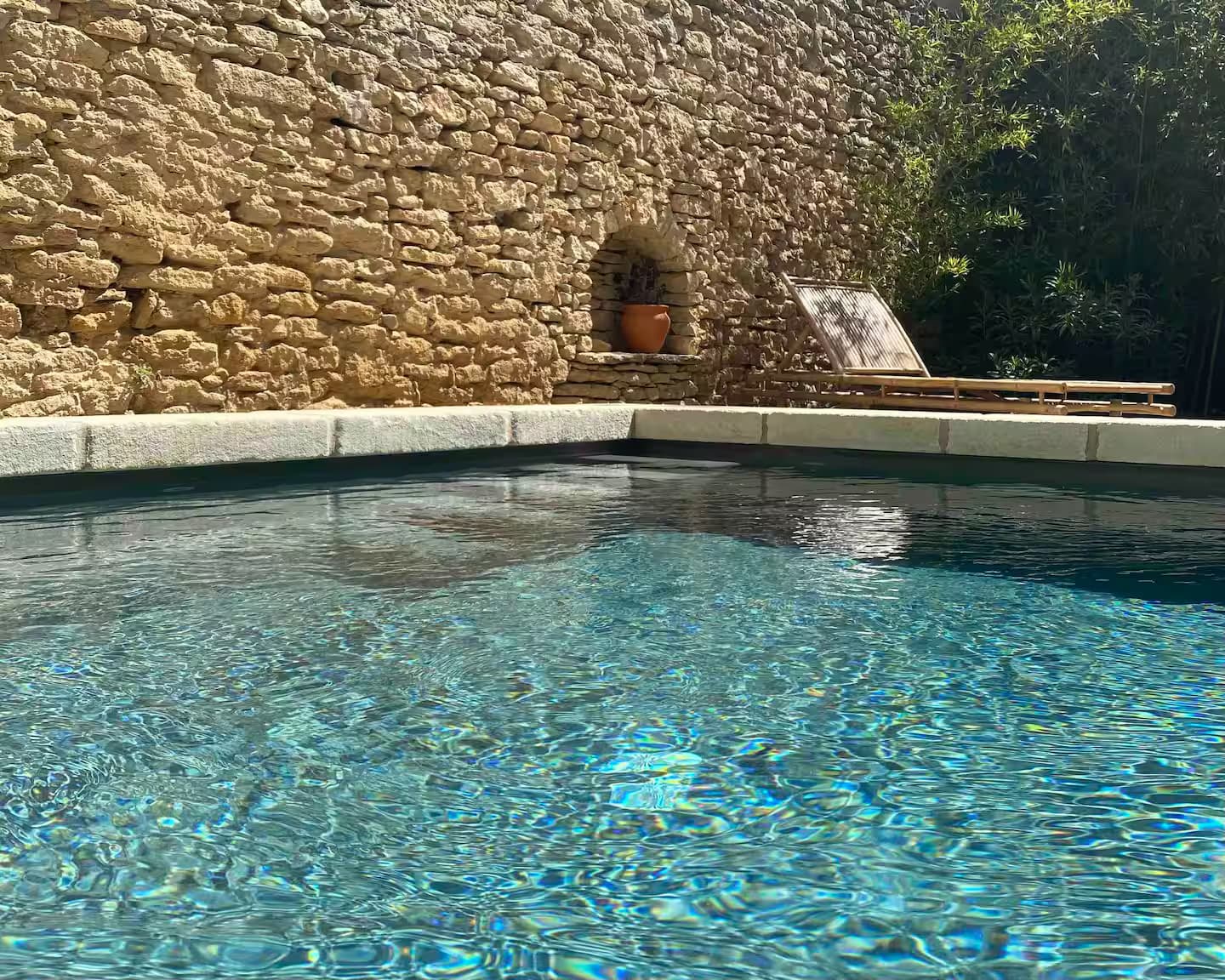 Une piscine bleu clair à côté d'un mur de pierre rustique, avec une chaise longue et une plante en pot en arrière-plan, partiellement ombragée par de la verdure.