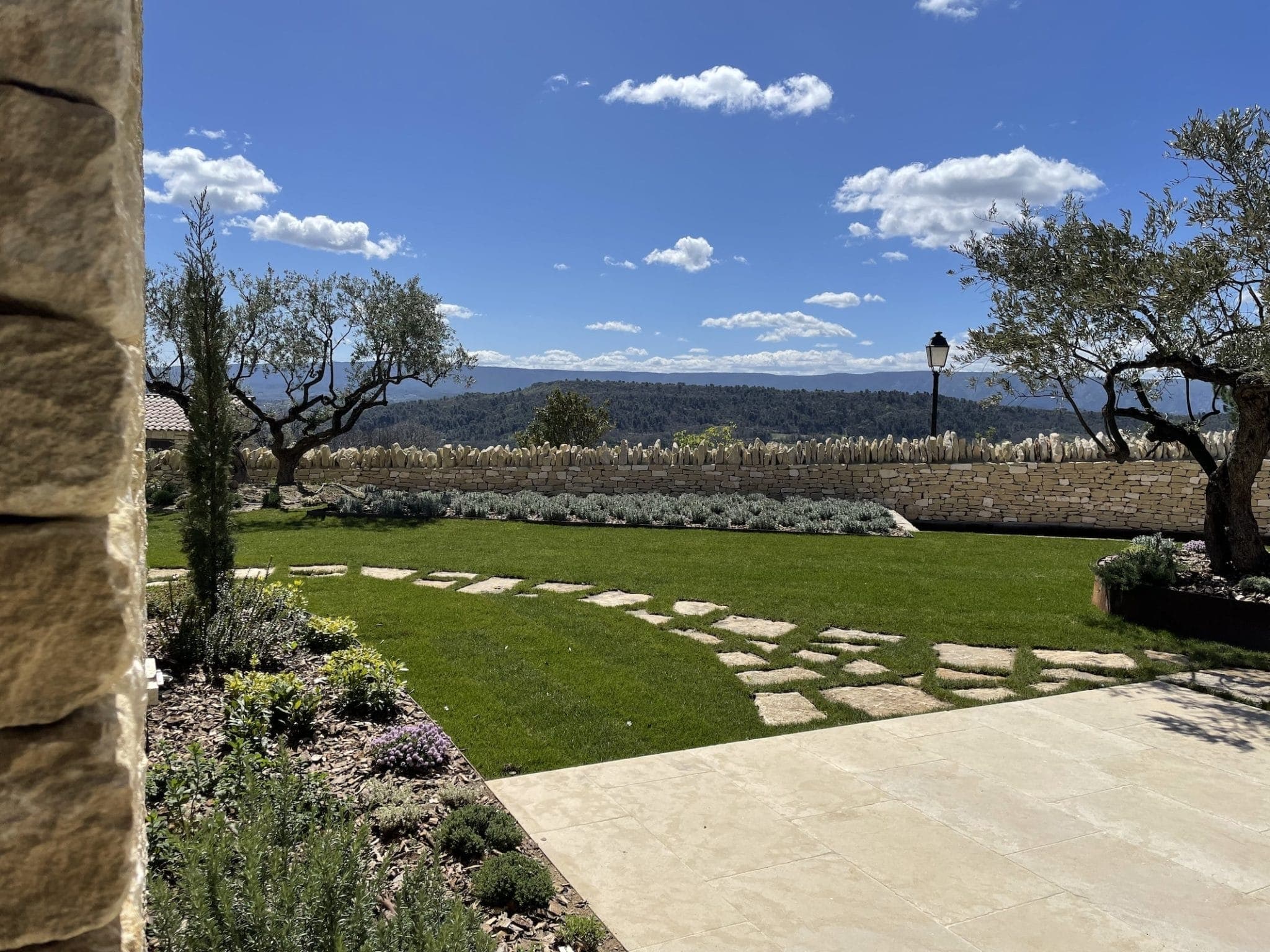 A sunlit garden features a neatly trimmed lawn with a stone path leading through it. Olive trees and shrubs adorn the landscape. A stone wall borders the garden, with hills and a clear blue sky in the background.