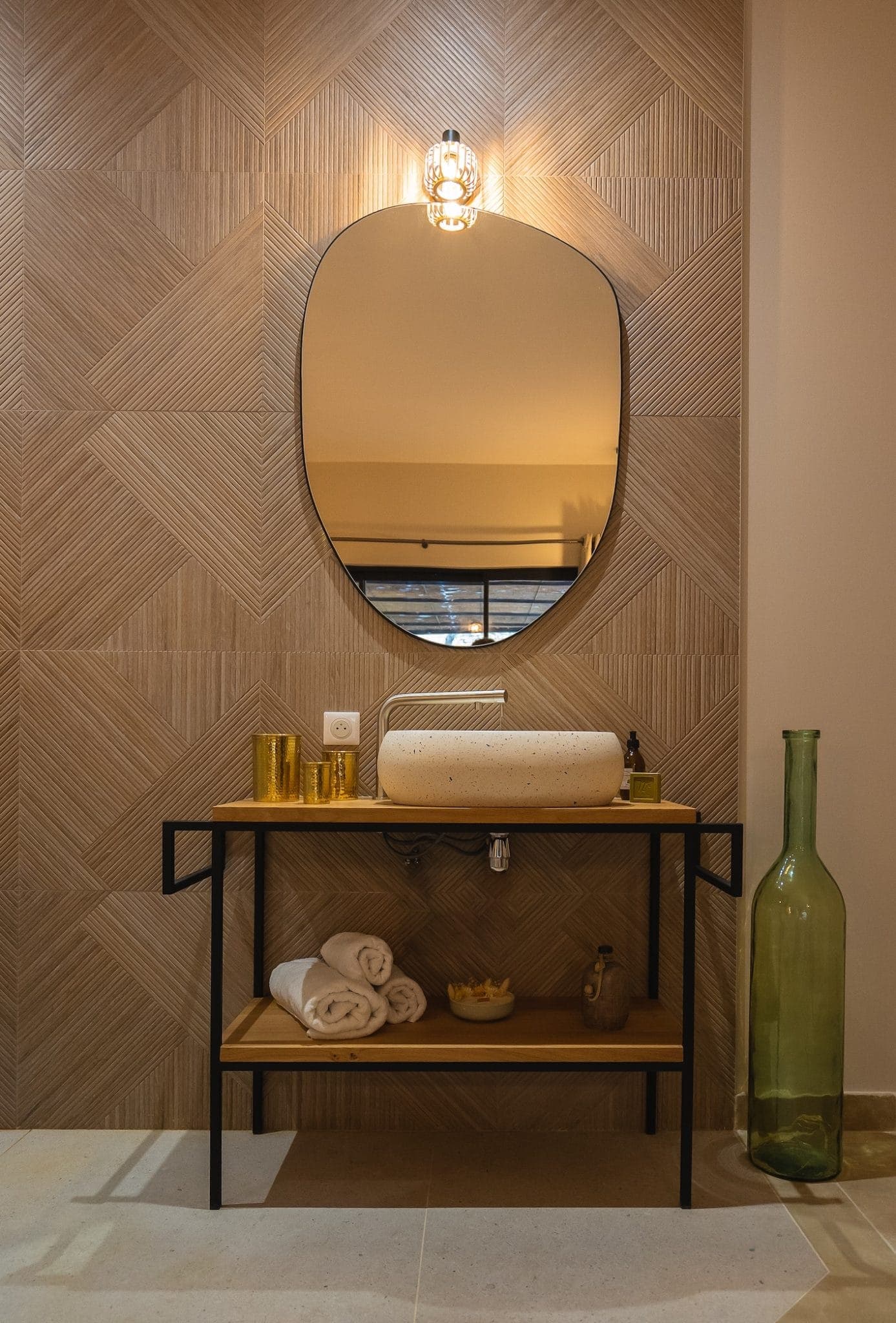 A modern bathroom with a geometric tile wall, oval mirror, and a stone sink on a wooden vanity with black frame. The vanity holds rolled towels and decor items. A tall green glass bottle vase stands on the floor, and a small light fixture is above the mirror.