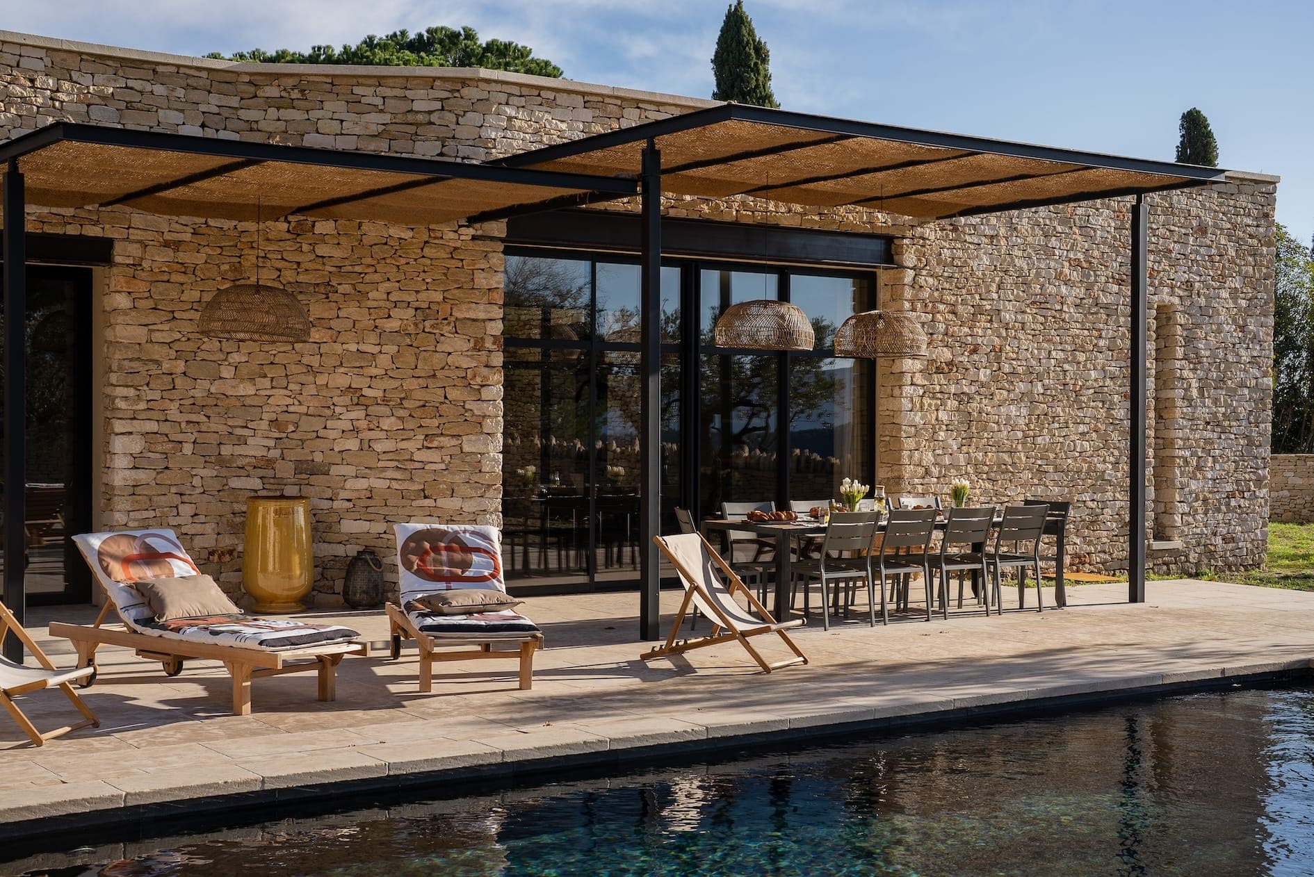 Un patio en pierre moderne comprend deux chaises longues en bois avec coussins, une grande table à manger extérieure avec chaises et une élégante pergola noire. L'espace donne sur une piscine rectangulaire, avec des arbres et un ciel clair en arrière-plan.