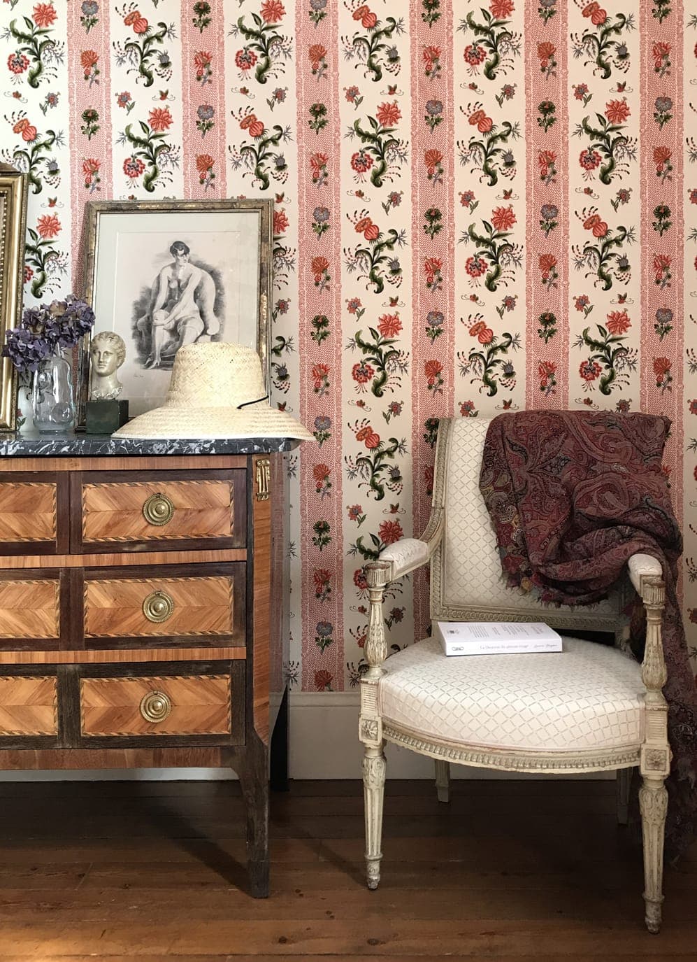 A vintage room with floral wallpaper features a wooden chest of drawers topped with a statue, straw hat and dried flowers. A white upholstered chair with a red-patterned shawl sits next to the dresser. A framed portrait leans against the wall.